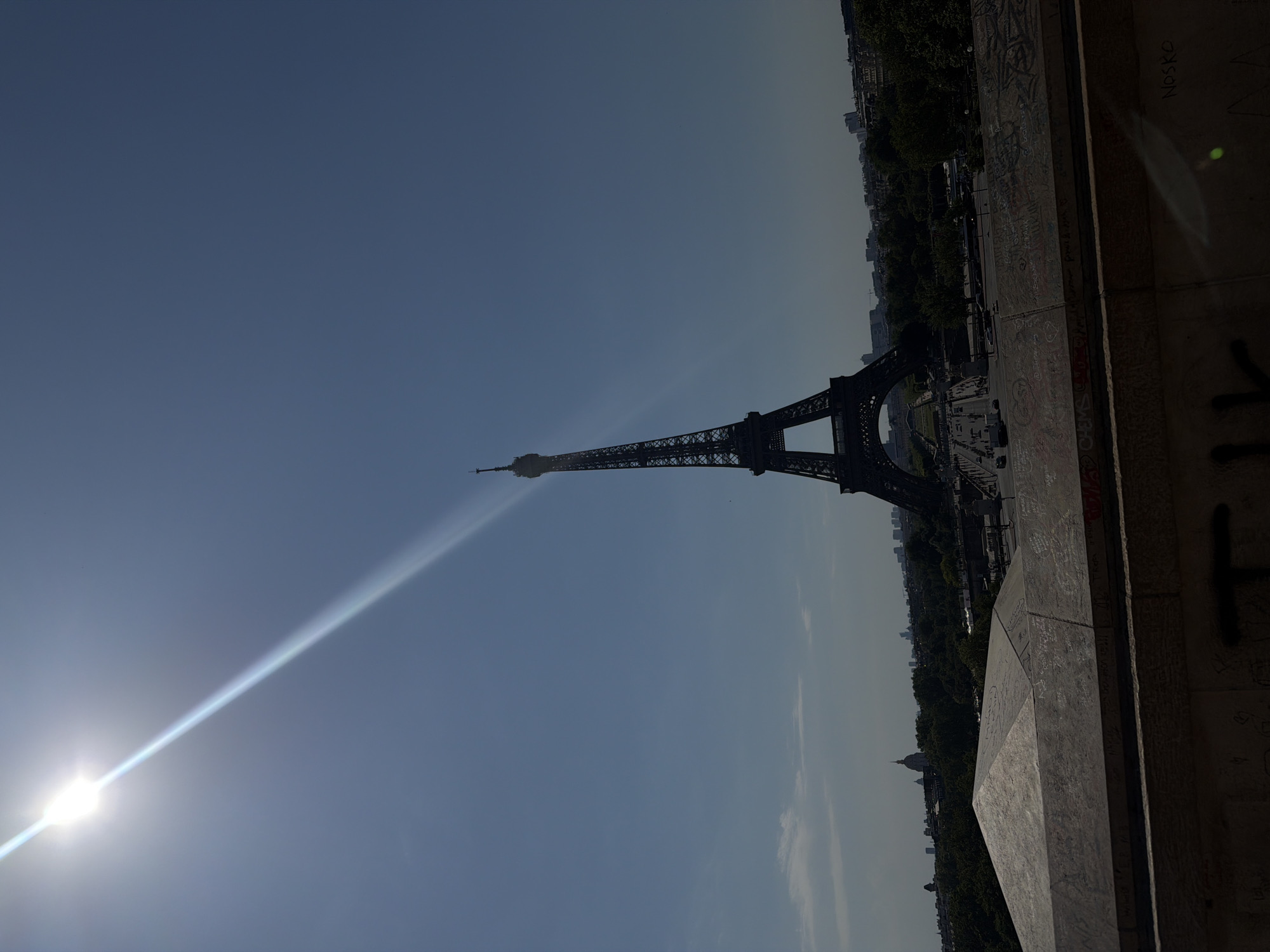 A bright sunbeam cuts diagonally across a clear blue sky, from the upper left towards the Eiffel Tower. The Eiffel Tower, a tall, dark lattice structure, stands prominently in the center-right of the image, silhouetted against the sky. The tower's base is visible, with a park or open area beneath it, where vehicles and people can be seen. In the foreground, a concrete structure, possibly a wall or a bridge railing, is covered in graffiti. The background shows a cityscape with trees and buildings, including a dome-shaped structure and distant skyscrapers. The overall impression is a daytime view of Paris, with the sun being a strong element in the composition.