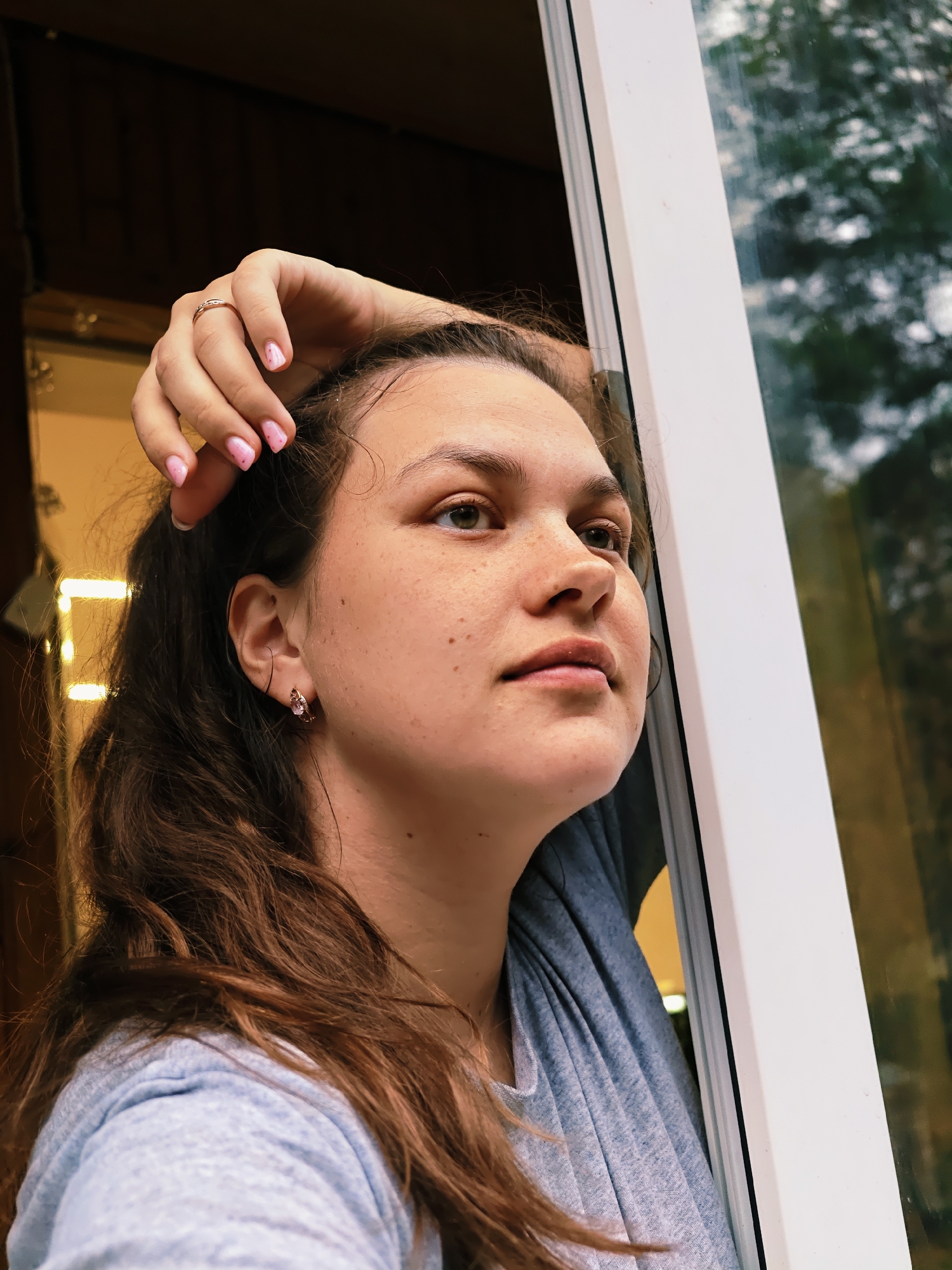 A fair-skinned woman with brown hair is looking out a window. She has freckles and is wearing a ring on her left ring finger and earrings. Her left hand rests on her head. The window has a white frame and reflects trees and sky. The woman is wearing a light blue shirt. The location is in Chaikovsky, Russia, based on other data.