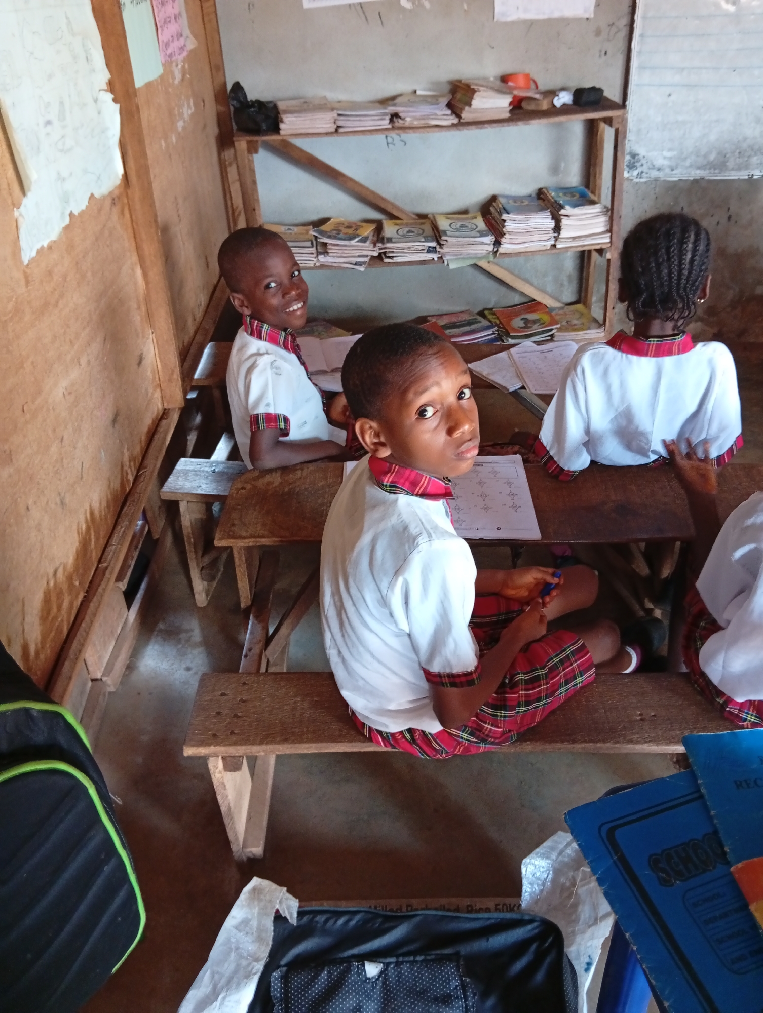 The image shows a classroom in Unknown, Unknown. Three children in school uniforms are seated at wooden desks, working on papers. A bookshelf with books is visible in the background. Two backpacks are on the floor.