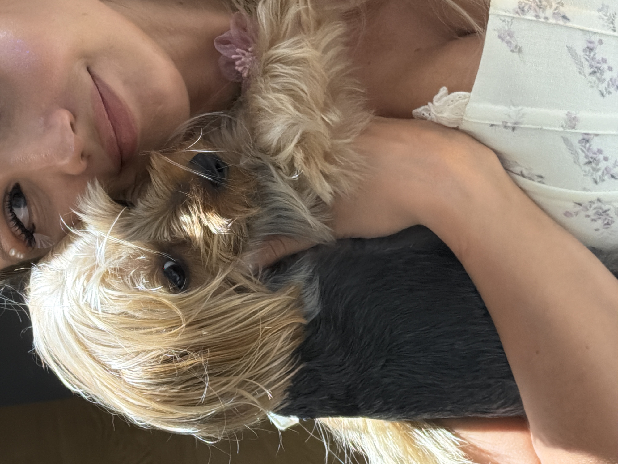 This media file features a close-up, overhead shot of a young woman lying down, holding a small, fluffy dog. The woman's face is partially visible, with her eye, cheek, and mouth in focus. She has fair skin and blonde hair. She is wearing a light-colored top with a floral pattern and delicate lace trim. A small pink flower adornment is visible in her hair. The dog, a Yorkshire Terrier, is nestled close to her, with its face obscured by its own blonde fur. Only one of its dark eyes is clearly visible. The background appears to be a warm, neutral color, possibly a bed or couch. The lighting suggests it is daytime, with a soft, natural light illuminating the scene. The overall mood is one of intimacy and affection between the woman and her pet. No text is visible in the image. The location context is Thep Krasatti, Thailand.