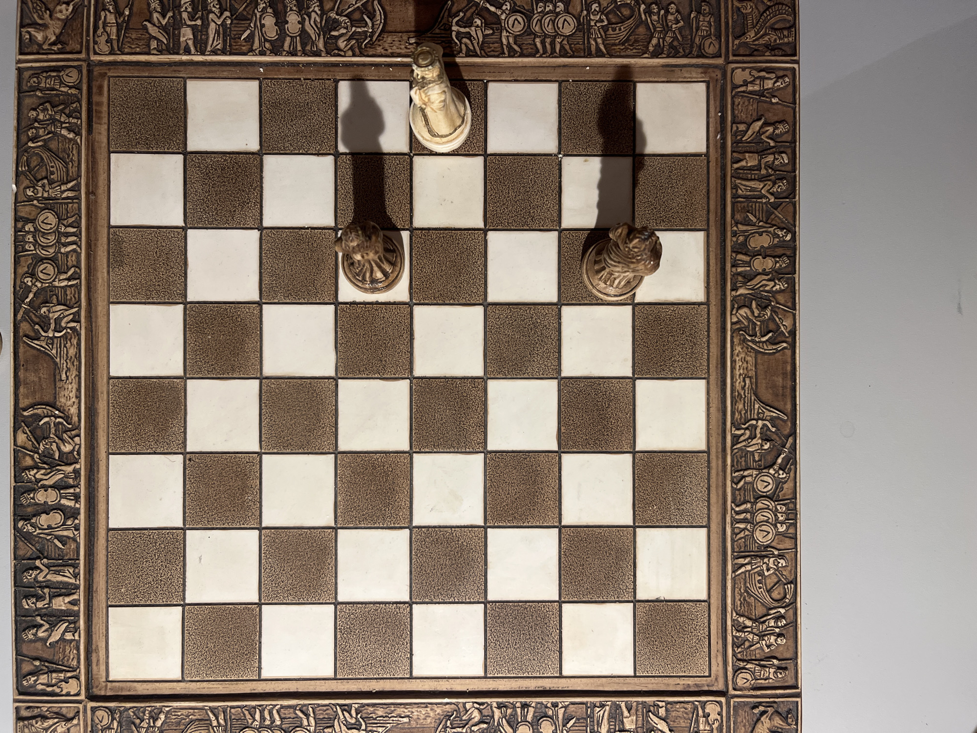 The image shows a close-up, top-down view of a chessboard with three chess pieces. The chessboard itself is intricately decorated with carved borders depicting figures and scenes, possibly of ancient warriors or mythological characters. The board has alternating light and dark squares.
There are two brown chess pieces, appearing to be knights, positioned on the board. One is on a dark square in the middle section of the board, and the other is on a dark square in the lower middle section. A single white chess piece, also appearing to be a knight, is positioned on a light square towards the right side of the board. The lighting suggests the board is placed on a flat surface under artificial light, casting shadows from the pieces. The surrounding environment is plain and out of focus, with a gray surface visible below the board.
There is no information in the image that would indicate its location in Minsk, Belarus.