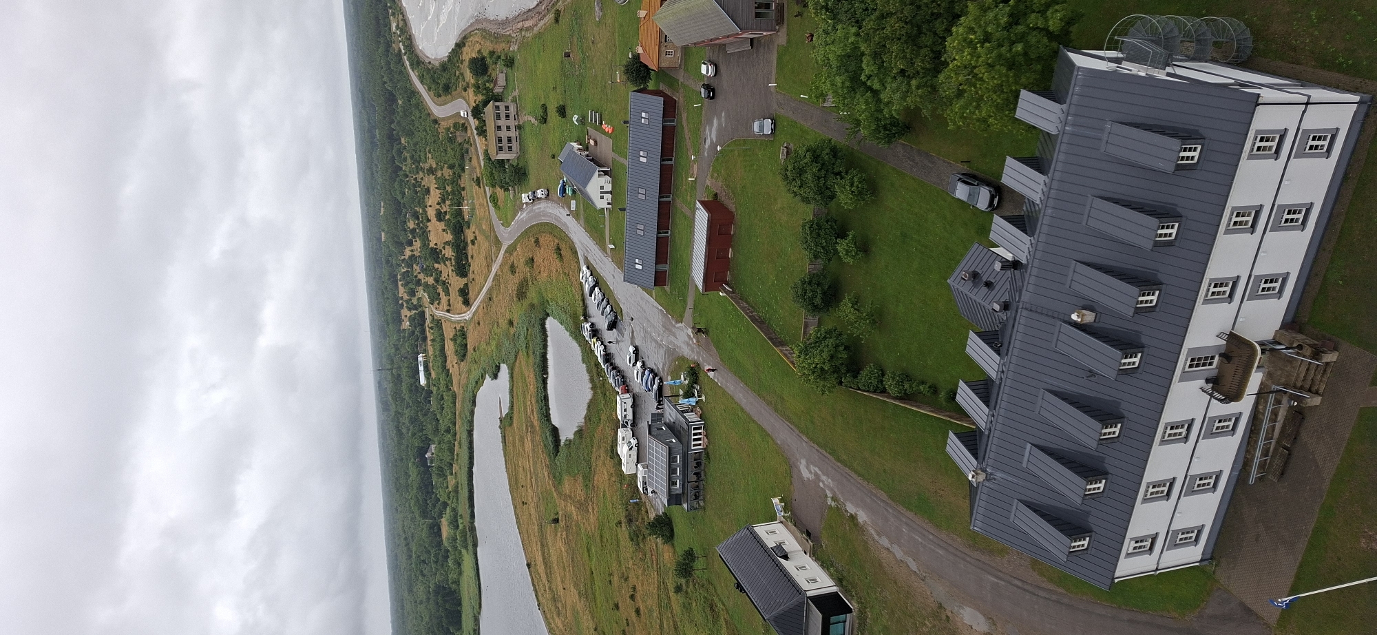 This aerial photograph depicts a coastal landscape in Sääre, Estonia, under an overcast sky. The scene features a body of water, possibly a bay or inlet, on the left, with a shoreline characterized by marshy vegetation and scattered trees. A road winds through the landscape, connecting various structures.
Prominent among the structures is a large, modern building with a dark gray, multi-pitched roof and white siding, featuring numerous windows. To the right of this building, a narrow, light-colored staircase with railings leads up to a platform. Further to the right, two large, circular metallic structures, resembling wind turbines or observation decks, are visible on top of another section of the building.
Scattered throughout the scene are smaller buildings, including red, rectangular structures and other modern-looking residences or apartments with dark roofs. A parking lot filled with several cars and what appear to be small boats or kayaks is situated along the road near some of these buildings. A few individuals can be seen scattered among the vehicles and buildings, suggesting activity and perhaps a sense of community or a public space. The overall impression is one of a developed coastal area with a blend of residential and potentially recreational facilities. The weather appears to be cloudy and potentially humid, typical of a coastal environment. No visible text is present in the image.