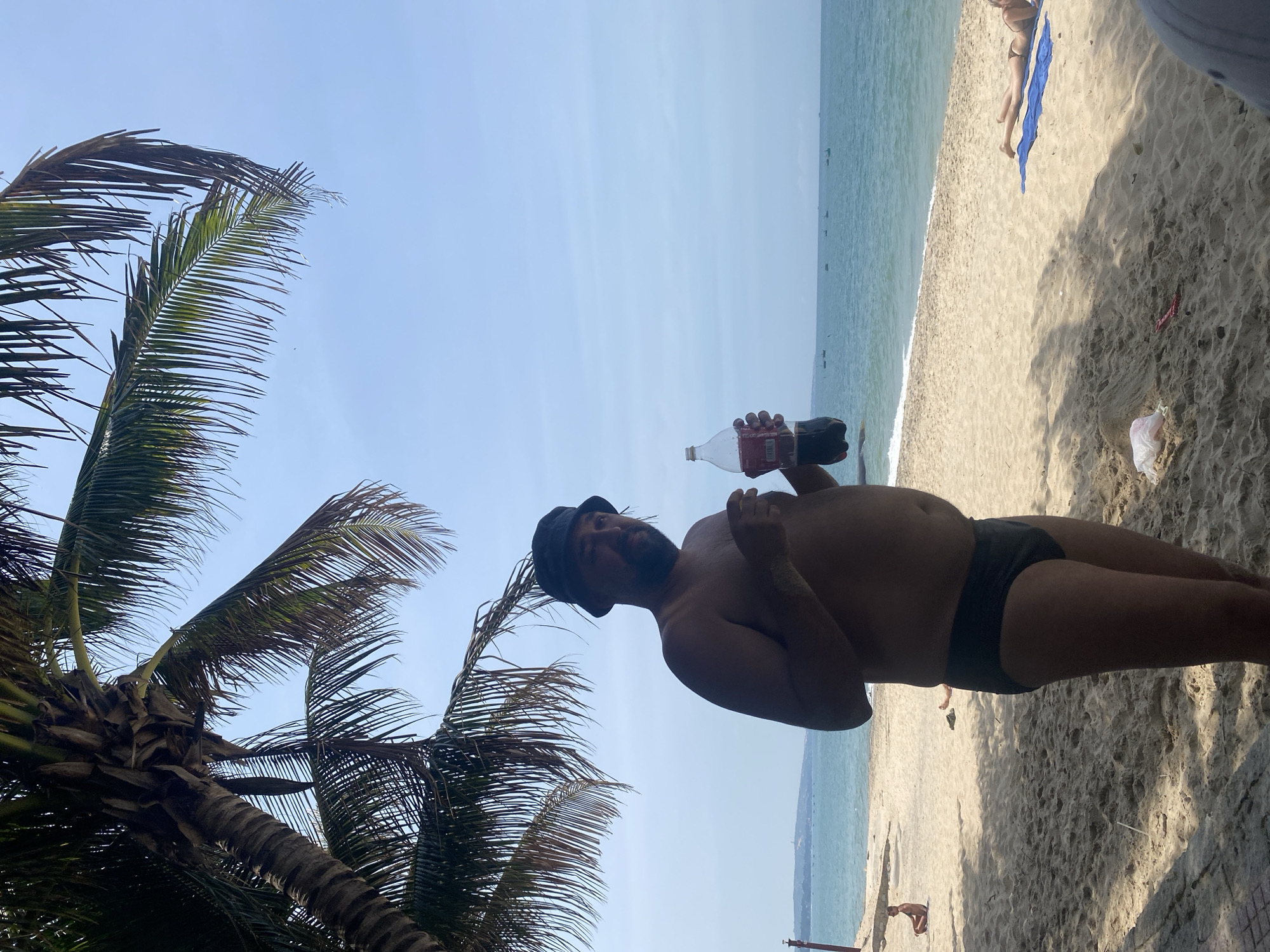 A man wearing a dark bucket hat and dark swim trunks stands on a sandy beach under the shade of a palm tree. He is holding a clear plastic bottle filled with a dark liquid, appearing to be Coca-Cola. The man is looking towards the viewer, and his left hand is slightly raised, holding what appears to be a small item. In the background, the ocean stretches to the horizon under a clear blue sky. Small fishing boats are visible on the water. The beach is sparsely populated with other individuals, including a person sitting in the distance and another person lying on a blue towel. The environment is a tropical beach setting.