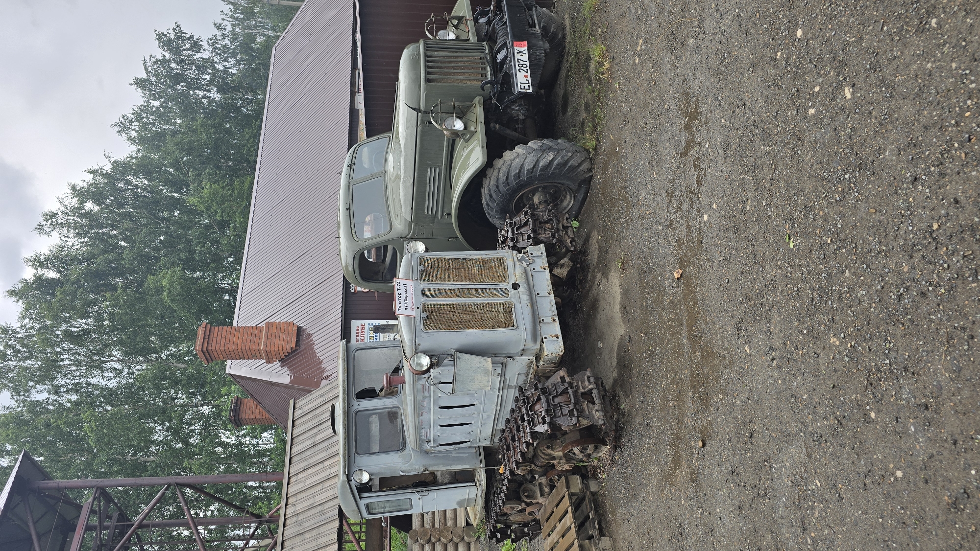 The image shows a dusty, outdoor scene with two vintage vehicles parked on gravel. In the foreground, a gray tracked tractor, possibly a DT-74 based on the sign, is positioned left of center. It has visible rust on its tracks and a bucket hanging from its front. To the right of the tractor, a green military-style truck is parked. The truck has a license plate that reads "EL 287 X". Behind the vehicles, a brown-roofed building with brick chimneys is visible, along with a metal tower structure to the far left. The background consists of a dense line of green trees under a cloudy sky. The environment appears to be a museum or an exhibition of historical machinery. The location is stated to be in the city of Bol'shoi Mashlyak, Russia. There are no people visible in the image.