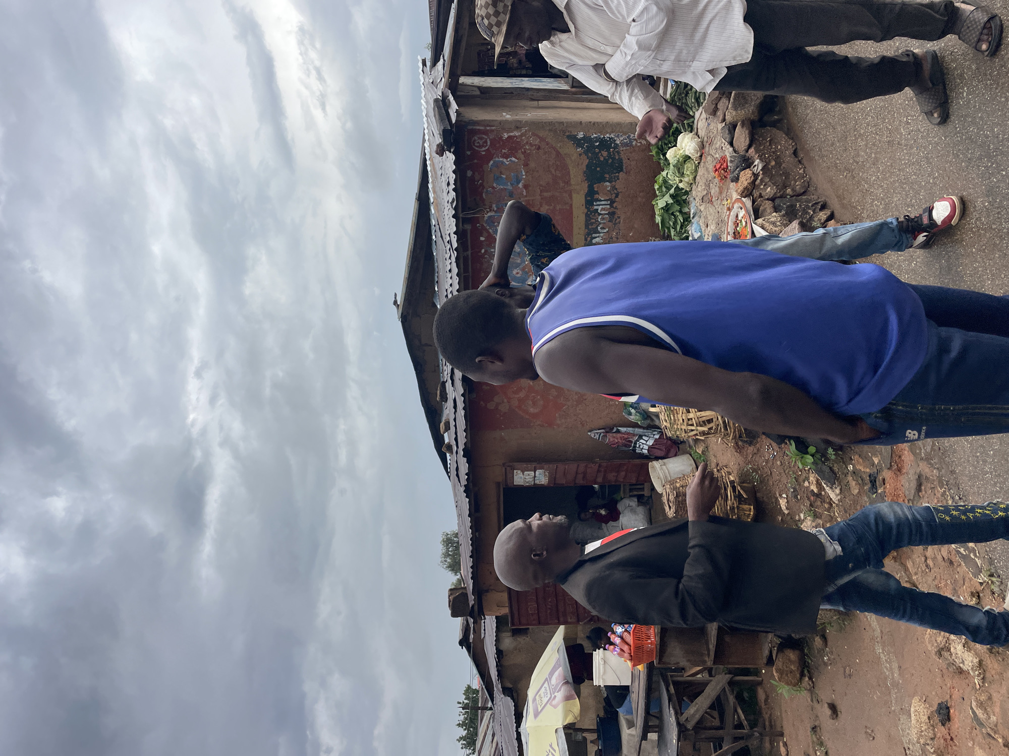 This image captures an outdoor scene, likely a bustling street market or commercial area in Jos, Nigeria, under an overcast sky.
In the foreground, a man is seen from behind, wearing a royal blue tank top. Adjacent to him, a bald man in a black jacket worn over a red shirt, blue jeans with distinctive yellow embroidery, and white sneakers, stands facing towards a building on the left side of the frame.
Behind these individuals, an old, weathered building with a corrugated iron roof serves as a backdrop. Its walls display faded, colourful paint and possibly remnants of advertisements or graffiti. Some text, possibly "OODL" and another word "POWER," is faintly discernible on the building's wall, though largely indistinct.
Near the building's entrance, market goods are displayed on the ground. A pile of green vegetables, resembling cabbage or lettuce, is visible, alongside smaller clusters of what appear to be red vegetables or spices. A man in a white striped shirt and a checkered cap stands near these goods, seemingly engaging in a transaction or survey. Another man, wearing a patterned dark shirt, leans against the building.
The ground is a mix of dirt and uneven patches, characteristic of a marketplace, with scattered baskets and containers. A yellow and white market umbrella or awning is partially visible on the left, further suggesting a market setup. The grey, cloudy sky indicates either an early morning, late afternoon, or generally overcast weather. The scene depicts typical daily life and commerce in an urban or semi-urban setting.