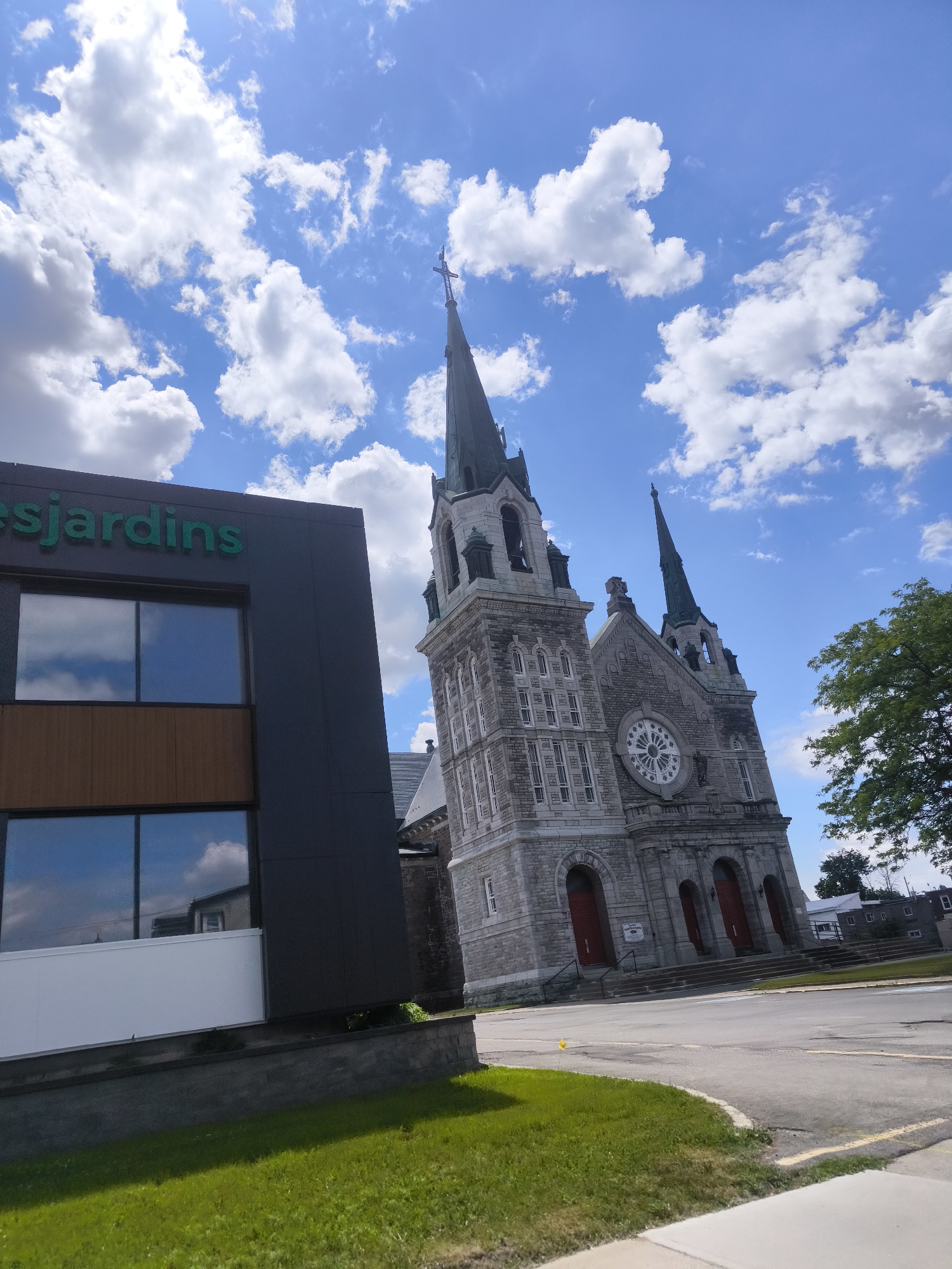 The image depicts a scene in Hawkesbury, Canada, focusing on a church and a modern building. The church, constructed of stone, features multiple spires, arched doorways with red doors, and a circular window. To the left of the church, a dark grey, modern building with large windows displays the green text "esjardins". The sky is blue with scattered white clouds, and a green tree is visible on the right. The ground in the foreground consists of green grass and asphalt pavement.
