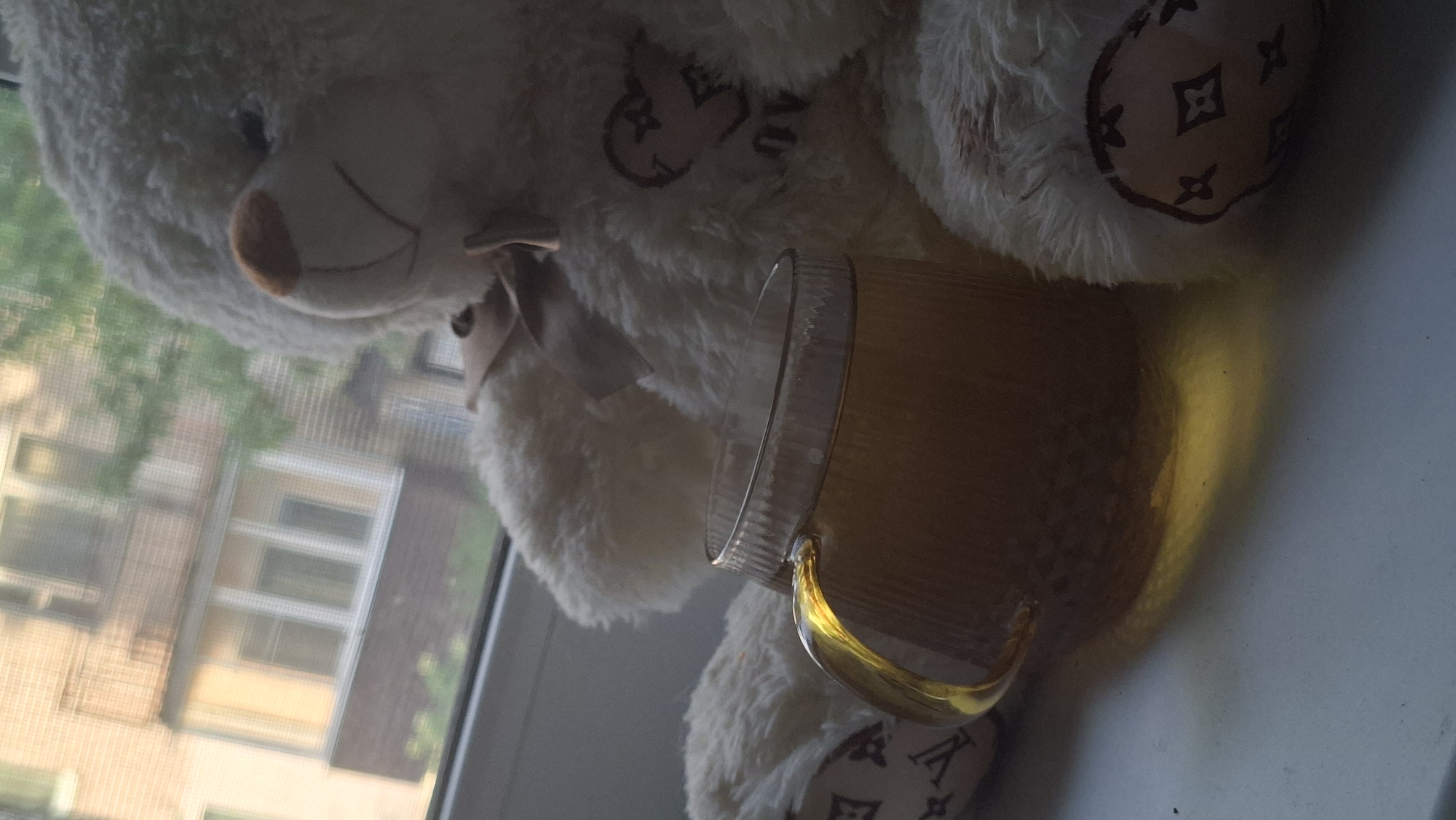 A fluffy white teddy bear sits on a windowsill, holding a glass mug of amber-colored liquid, possibly tea. The mug has a ribbed texture and a golden sheen where the liquid catches the light. The teddy bear has a brown bow at its neck and a stitched heart design on its chest. Its paws have a pattern resembling the Louis Vuitton monogram. Outside the window, a brick building with balconies and trees can be seen, suggesting an urban environment. The lighting indicates it may be daytime, with sunlight casting shadows and highlighting the scene. This appears to be a cozy indoor setting, possibly a home in the city of Myadzyel, Belarus.