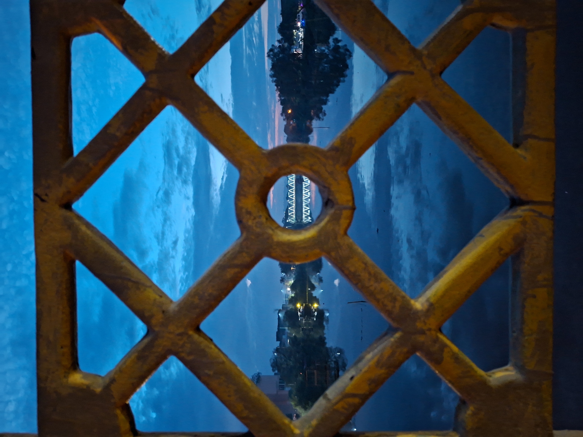 A photograph taken at dusk through a yellow, diamond-shaped metal railing shows a serene scene of the Krueng Cunda river in Lhokseumawe, Indonesia. The railing's lattice work, with a central circular opening, frames a view of the tranquil water reflecting the twilight sky. The sky is a deep blue, streaked with wispy clouds, and the setting sun casts a faint glow on the horizon. On the far bank of the river, trees and buildings are visible, illuminated by scattered lights. A pedestrian bridge, constructed with a truss design, spans the river in the distance, its white structure stark against the darkening sky. The overall impression is one of quiet beauty and calm, typical of an evening in this coastal city.