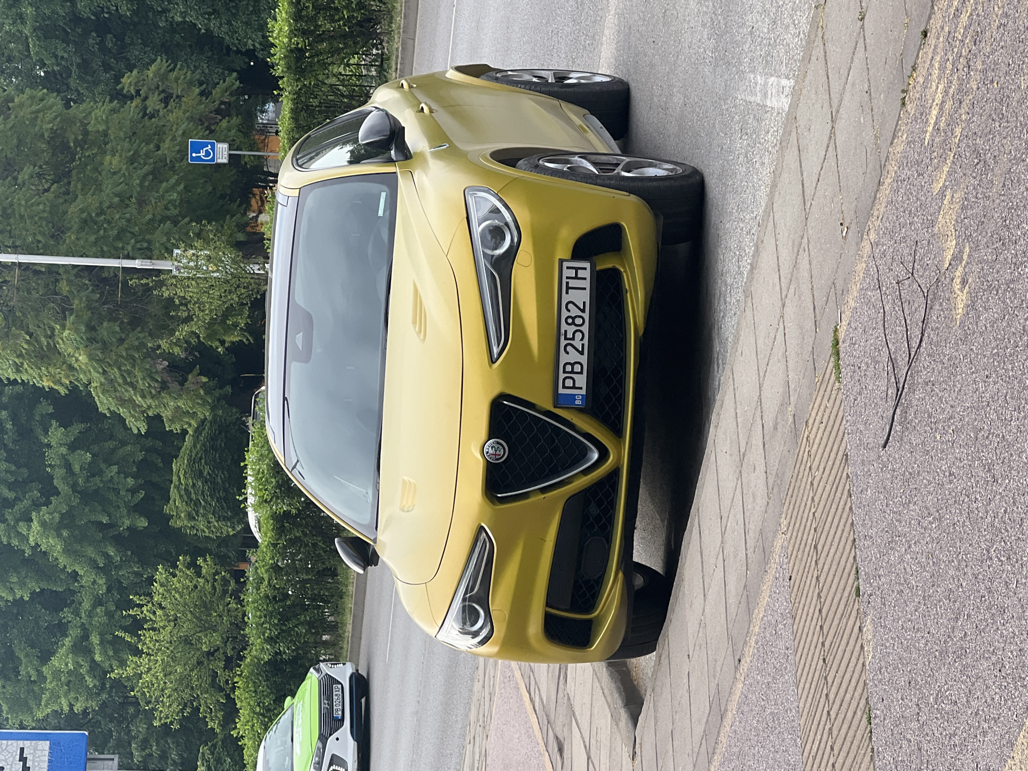 A yellow Alfa Romeo Stelvio SUV is parked on the side of a street in Plovdiv, Bulgaria. The car has a Bulgarian license plate that reads "PB 2582 TH". The vehicle is positioned on a sidewalk next to a road, with a line of green bushes and trees in the background. There is also a white car with a green stripe and a Bulgarian license plate in the distance. A blue sign with a wheelchair symbol is visible on a pole in the background. The sky is overcast.