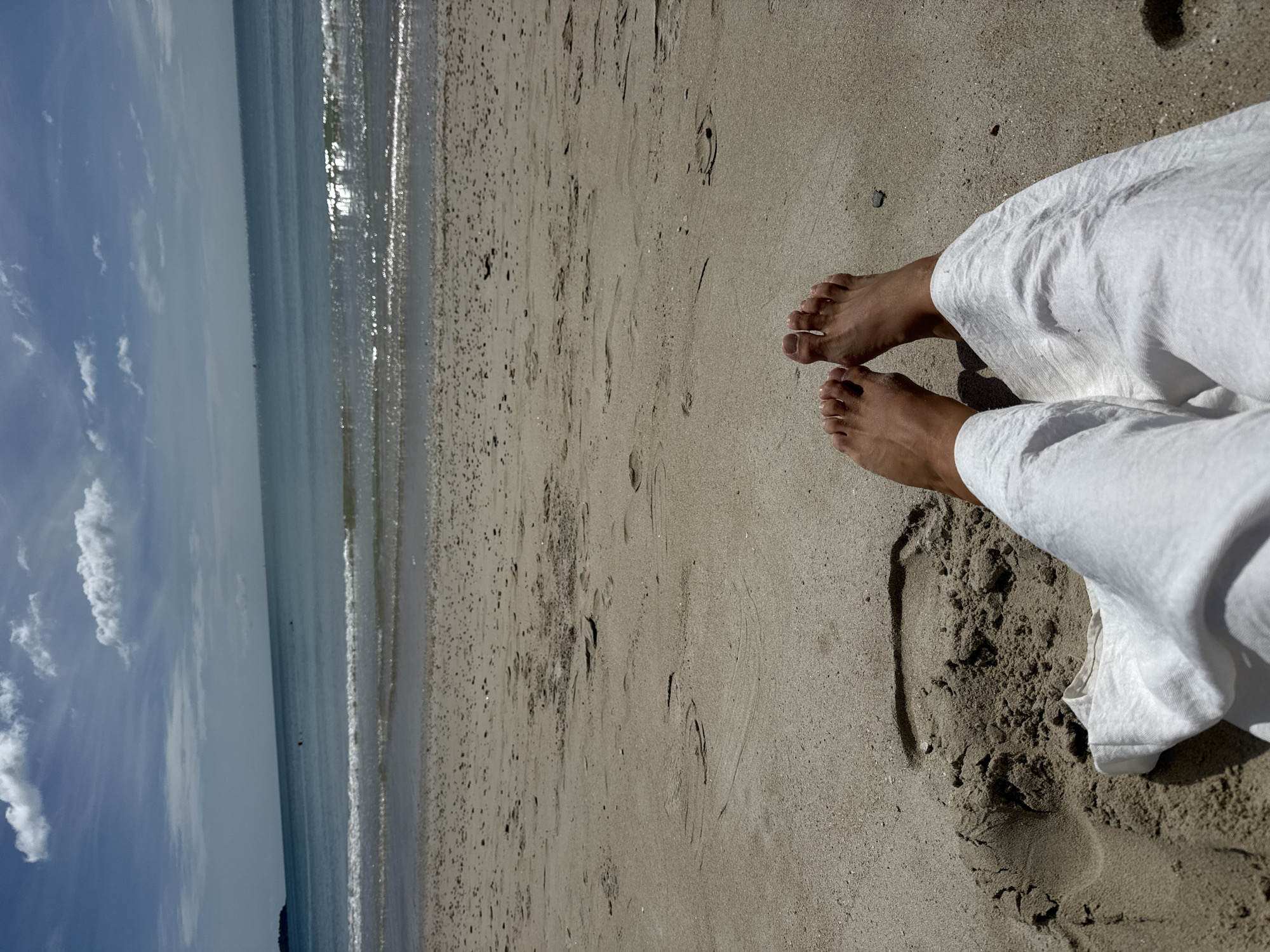 The image depicts a close-up view of a person's bare feet and white linen pants resting on a sandy beach in Ko Samui District, Thailand. The foreground is dominated by the sand, with visible footprints and a large indent in the sand, possibly from where the person was sitting or lying down. The person's feet are positioned centrally in the frame, with toes pointed upwards. Beyond the sand, the turquoise ocean stretches to the horizon under a partially cloudy blue sky. Gentle waves are breaking on the shore, creating a white foam line. In the distance, a faint silhouette of land is visible on the left side of the frame. The overall scene conveys a sense of relaxation and a tropical beach setting.