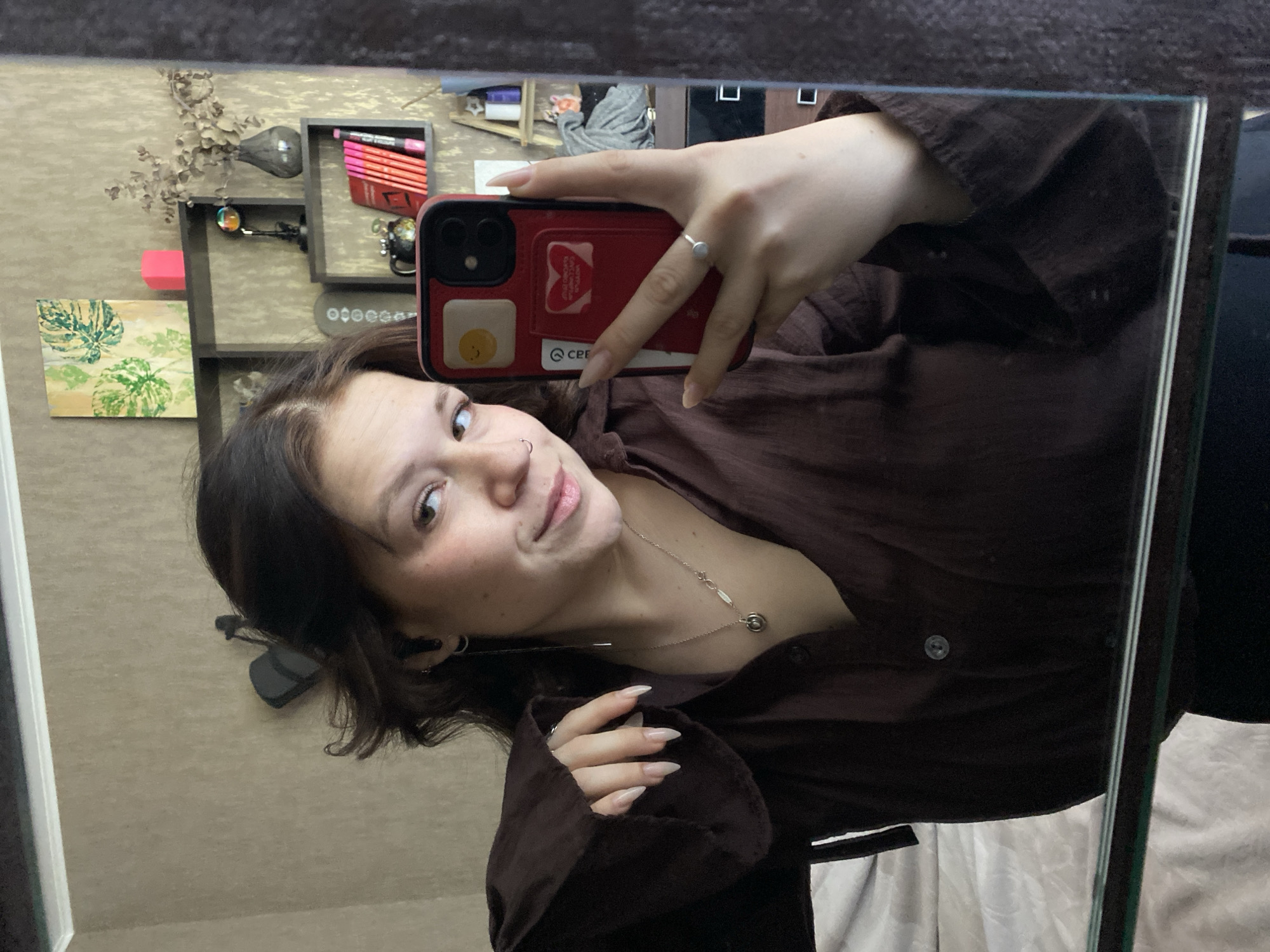 A woman is taking a selfie in a mirror. She is holding a red phone with a white credit card case on the back and a smiley face sticker on the phone case. The woman is wearing a dark brown button up shirt and a silver necklace. Her nails are long and painted light pink. She has brown hair and a nose ring. A silver ring is on her left hand ring finger. Behind her in the reflection is a wall with shelves holding pens, a small globe, jewelry, and a framed painting. The background suggests an interior space, potentially in a residential setting. The city and country are not definitively discernible from the image.