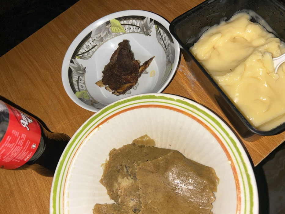The image displays a meal arranged on a wooden table, likely in an indoor setting in Debasco, Nigeria. In the upper left, a large portion of light brown, dense food, consistent with a swallow food like Amala or Eba, is contained within a white ceramic bowl featuring green and orange stripes on its rim. To the right of this bowl, a smaller white ceramic bowl, decorated with a grey and black leaf pattern, holds several pieces of dark brown, roasted meat, possibly ribs. At the top of the frame, a dark beverage in a clear plastic bottle with a prominent red label, partially visible as "Amigo" and indicating "35 CL," rests on the table. In the lower right corner, a black rectangular plastic container holds a pale yellow, semi-liquid substance, resembling custard or pap, with a small white plastic spoon inserted into it. No individuals are visible.