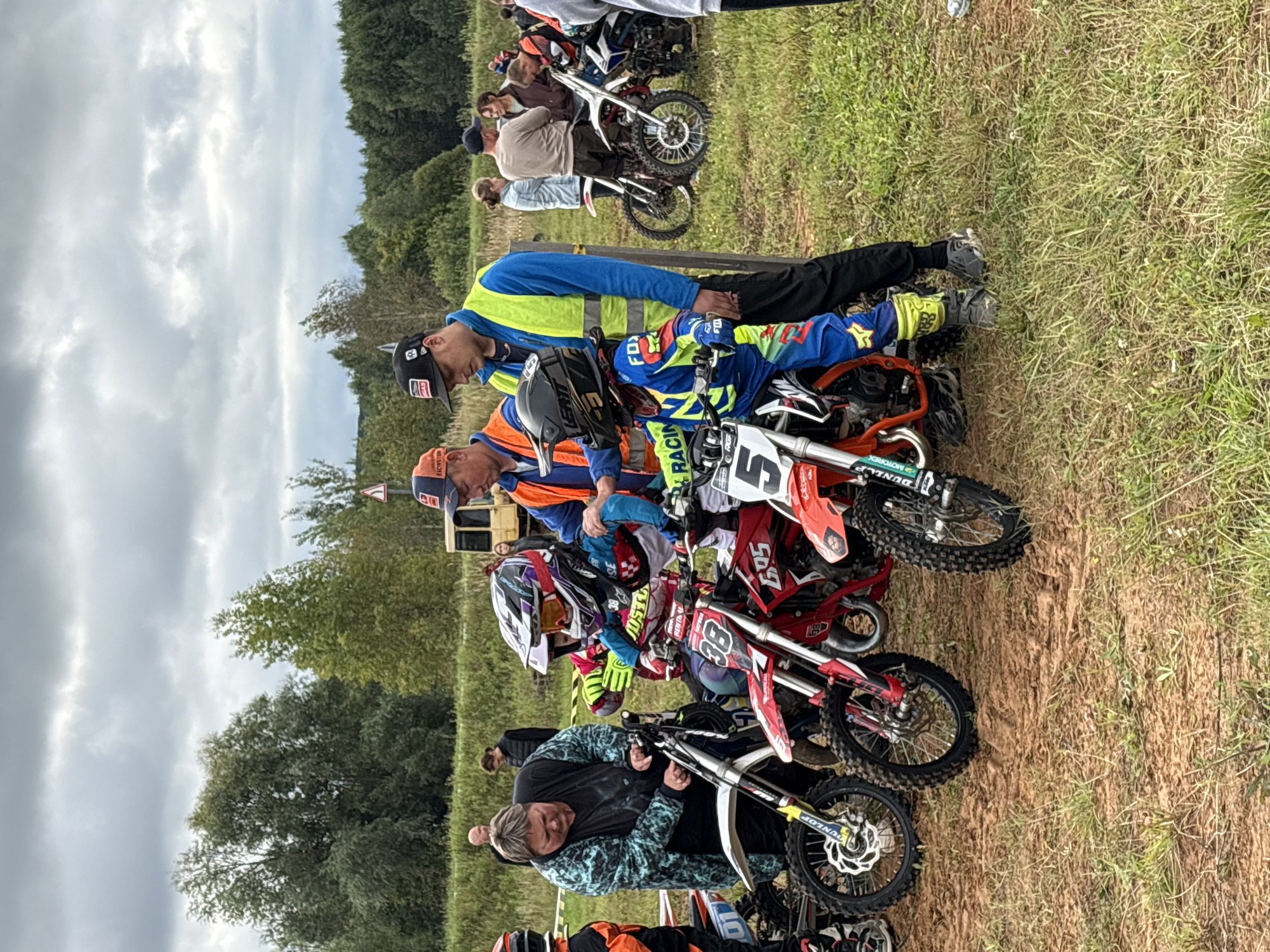 This image depicts a group of people, primarily children in motocross gear and adults, gathered around dirt bikes in an outdoor, grassy environment. The scene suggests a motocross event or practice session. In the foreground, three children are visible on or near their motorcycles, wearing full protective gear including helmets, goggles, and colorful racing suits. Adults are present, some appearing to be coaches or supervisors, assisting the children and the motorcycles. In the background, trees and a cloudy sky are visible, indicating an outdoor setting. The ground appears to be a mix of dirt and grass. The location of Балаки, Russia is not discernable from the image itself, but the scene is consistent with a rural or suburban area where such events might take place.