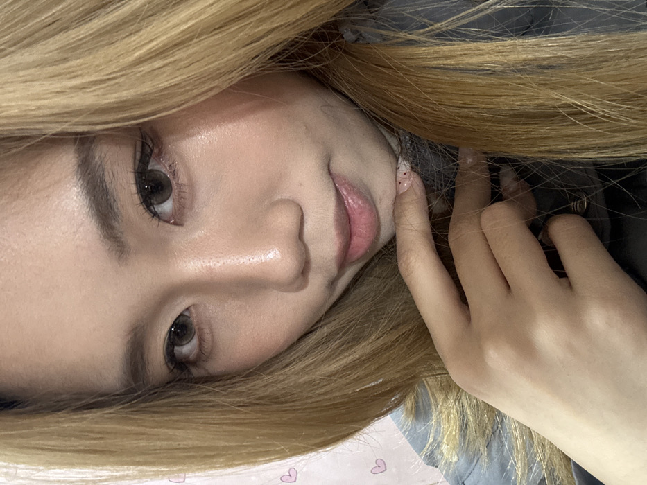 This close-up portrait depicts an East Asian female in an indoor setting, presumed to be within Shapingba District, China. The subject has long, straight, honey-blonde hair, parted slightly to the left, framing her face. Her complexion is fair with a luminous quality. She possesses large, dark brown eyes, possibly augmented by colored contact lenses, defined by prominent eyeliner and eyelashes. Her eyebrows are well-groomed. Her lips are subtly parted, displaying a pink tint. Her left hand is raised, with fingers lightly touching the area beneath her chin. The nails on her visible fingers are manicured with a light pink polish featuring small red/pink glitter accents. She is wearing a dark grey, high-collared garment. The background is out of focus, appearing as a light pink surface adorned with small, indistinct pink heart graphics.