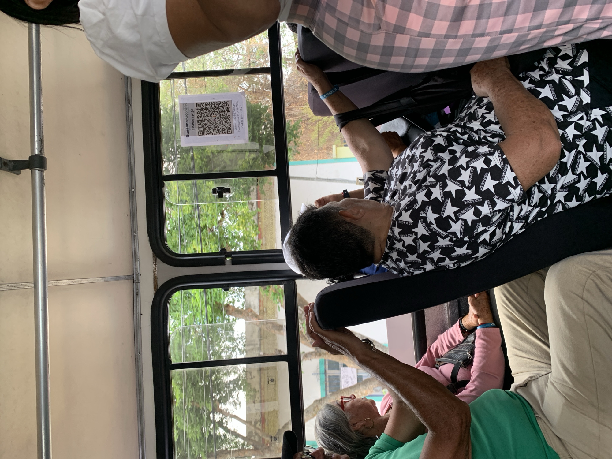 The image shows people riding in a bus, as evidenced by the windows, seats, and handrails. A sign in the window shows a QR code and states "Banesco PagoMóvil," "Escanea y paga," a phone number "0424-8240684," an identification number "V-14685036," and bank code "0134." One person is wearing a shirt with a repeating star and "GOLDEN GOOSE" pattern. Another person is wearing a green shirt. A bracelet on one person's wrist reads "ALTAGRACIA." The QR code sign suggests the location is in Venezuela. The sign mentions bank information, which leads to the conclusion that the image was taken in Santa Ana, Venezuela.