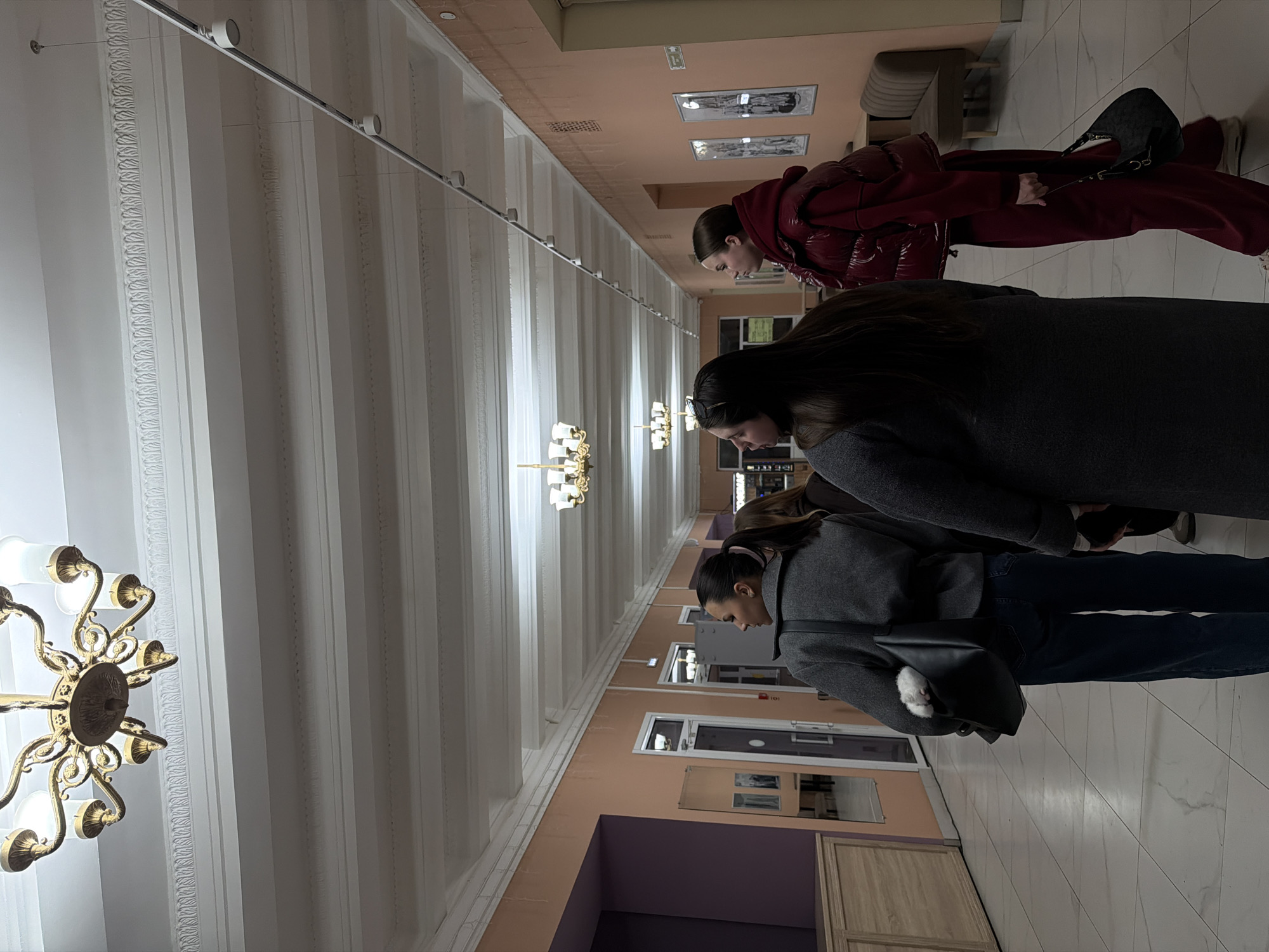 The image shows the interior of a building in Perm, Russia. The building has a long hallway with a high ceiling decorated with chandeliers and ornate moldings. The walls are painted in shades of peach and purple, and there are doorways and framed pictures along the walls.
Three women are visible in the hallway. One is wearing a red puffer jacket and dark pants. Another is wearing a gray coat. The third is wearing a gray jacket and blue jeans. She has a black bag on her side which contains a white animal. The women appear to be looking at something.
The floor is tiled in a light color with a grid pattern. There are also some pieces of furniture in the hallway, including benches.