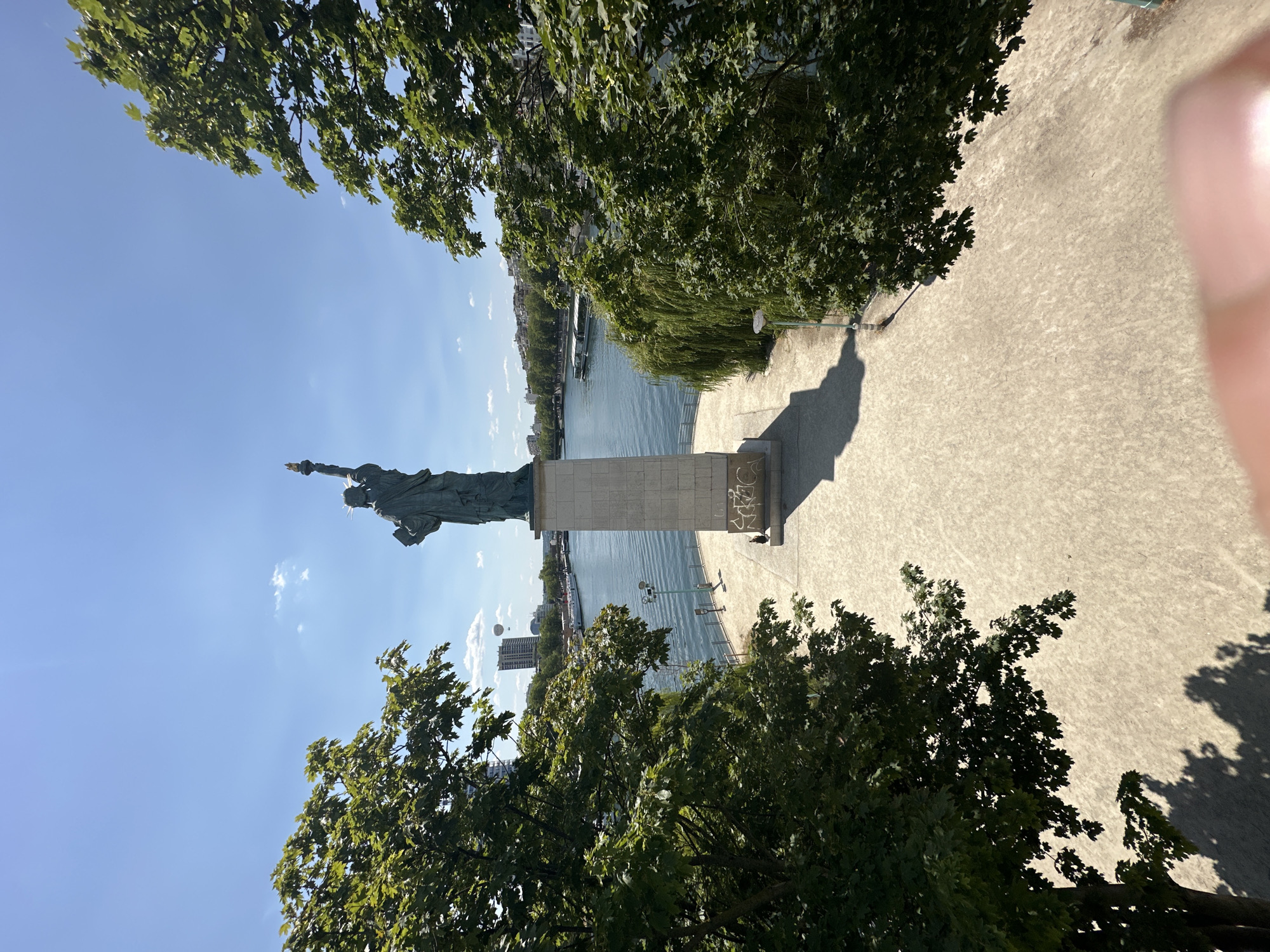 The image depicts the "Statue of Liberty" on the Île aux Cygnes in Paris, France. The statue, a smaller replica of the original in New York, stands on a stone pedestal overlooking the Seine River. A riverboat is visible on the water, and buildings line the opposite bank. Lush green trees frame the scene on both sides, casting shadows on a paved walkway leading to the statue. The sky is clear blue with a few scattered clouds. There is one person standing on the walkway near the base of the statue, and a dog is also visible. The overall impression is a sunny day in a park-like setting by the river.