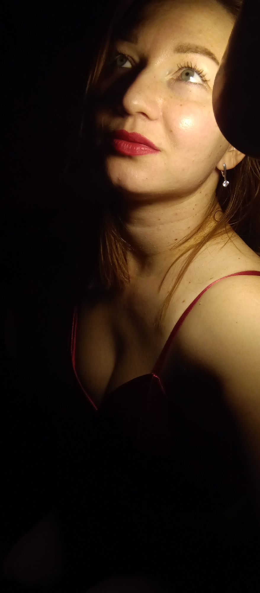 A woman is captured in a low-light, close-up shot. She has fair skin, red lipstick, and is wearing a red dress with thin straps. The lighting is uneven, with the left side of her face and body obscured in darkness, and the right side highlighted by a strong, directed light source. She has an earring on her right ear. The background is dark. The location is unknown.