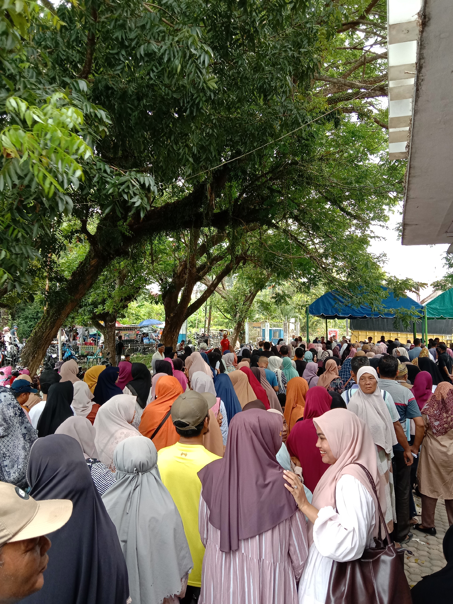 A large crowd of people, mostly women, is gathered in an outdoor location in Lhokseumawe, Indonesia. Many of the women are wearing hijabs of various colors, including gray, orange, purple, and pink. Several men are also present, some wearing hats. The crowd appears to be waiting in line, possibly for some kind of event or service. In the background, there are tents with blue and green covers. A large tree provides shade over a portion of the crowd. The location appears to be near a building with a concrete facade. Some motorcycles and bicycles are visible in the background.
