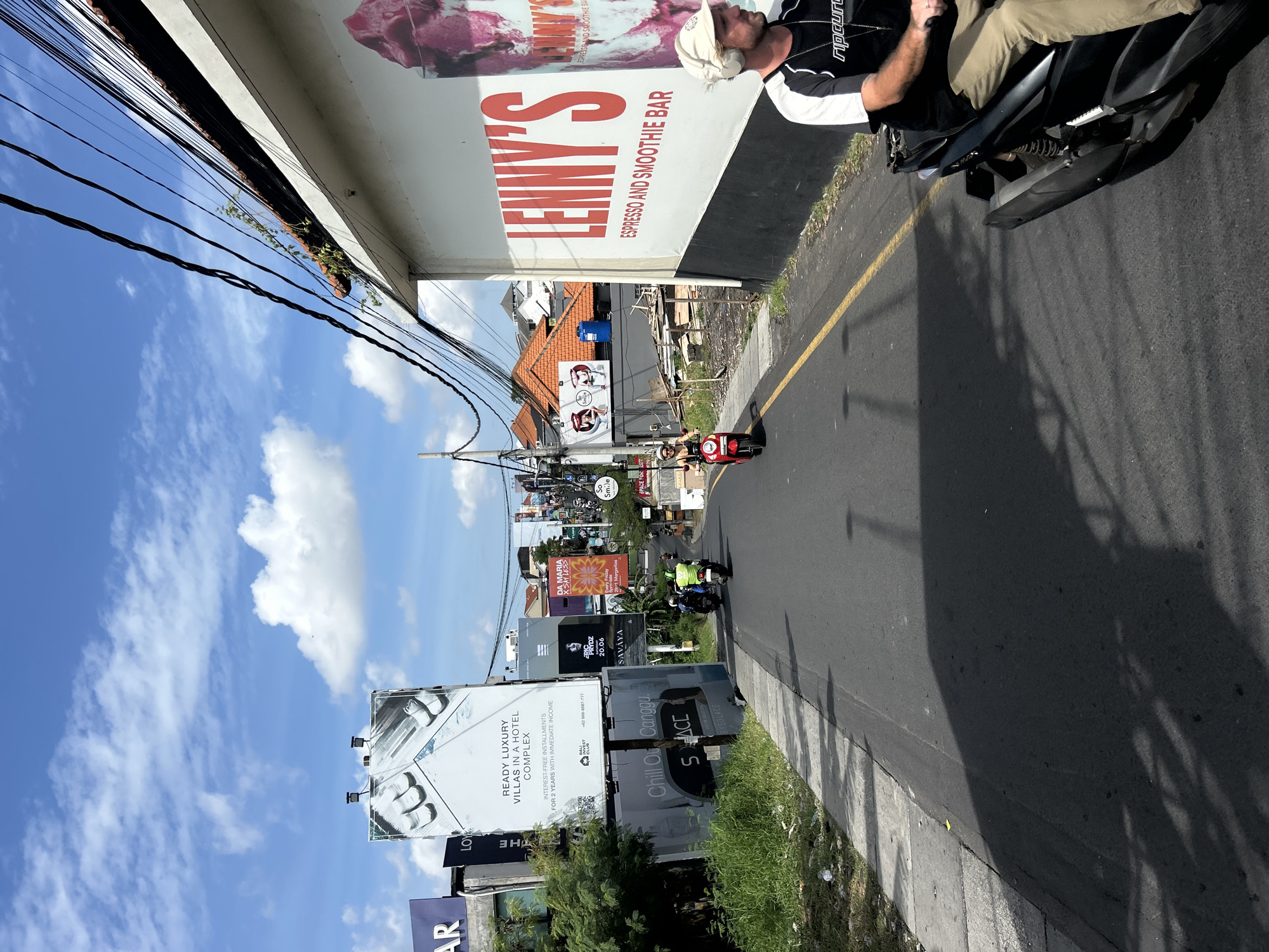 The image shows a street scene in North Kuta, Indonesia, on a bright, sunny day with a blue sky and scattered white clouds. The perspective is from a moving vehicle, looking down a paved road lined with buildings and billboards.
On the left side of the road, there are several large billboards. One prominent billboard displays an advertisement for "READY LUXURY VILLAS IN A HOTEL COMPLEX" with "INTEREST-FREE INSTALLMENTS FOR 2 YEARS WITH IMMEDIATE INCOME." Below this, another billboard reads "Chill Out Canggu S SPACE" and has a logo. Further down the road on the left, there's a billboard with a partial view of "AR."
On the right side of the road, a large white building features a prominent red sign that reads "LENNY'S ESPRESSO AND SMOOTHIE BAR." A portion of an advertisement showing a pink dessert is also visible on this building.
Several people are visible on motorbikes. In the mid-ground, a person in a bright green vest is riding a motorbike. To the right of that, a person on a red motorbike is also visible. In the foreground on the right, a man wearing a white cap and headphones is riding a motorbike, his torso and legs visible as he looks ahead.
The environment appears to be a developing or busy commercial area with overhead electrical wires strung across the street. Vegetation is present along the sides of the road, including some trees and overgrown grass. The road itself has a yellow dividing line. Shadows of vehicles and objects are cast on the road surface, indicating the direction of the sun.