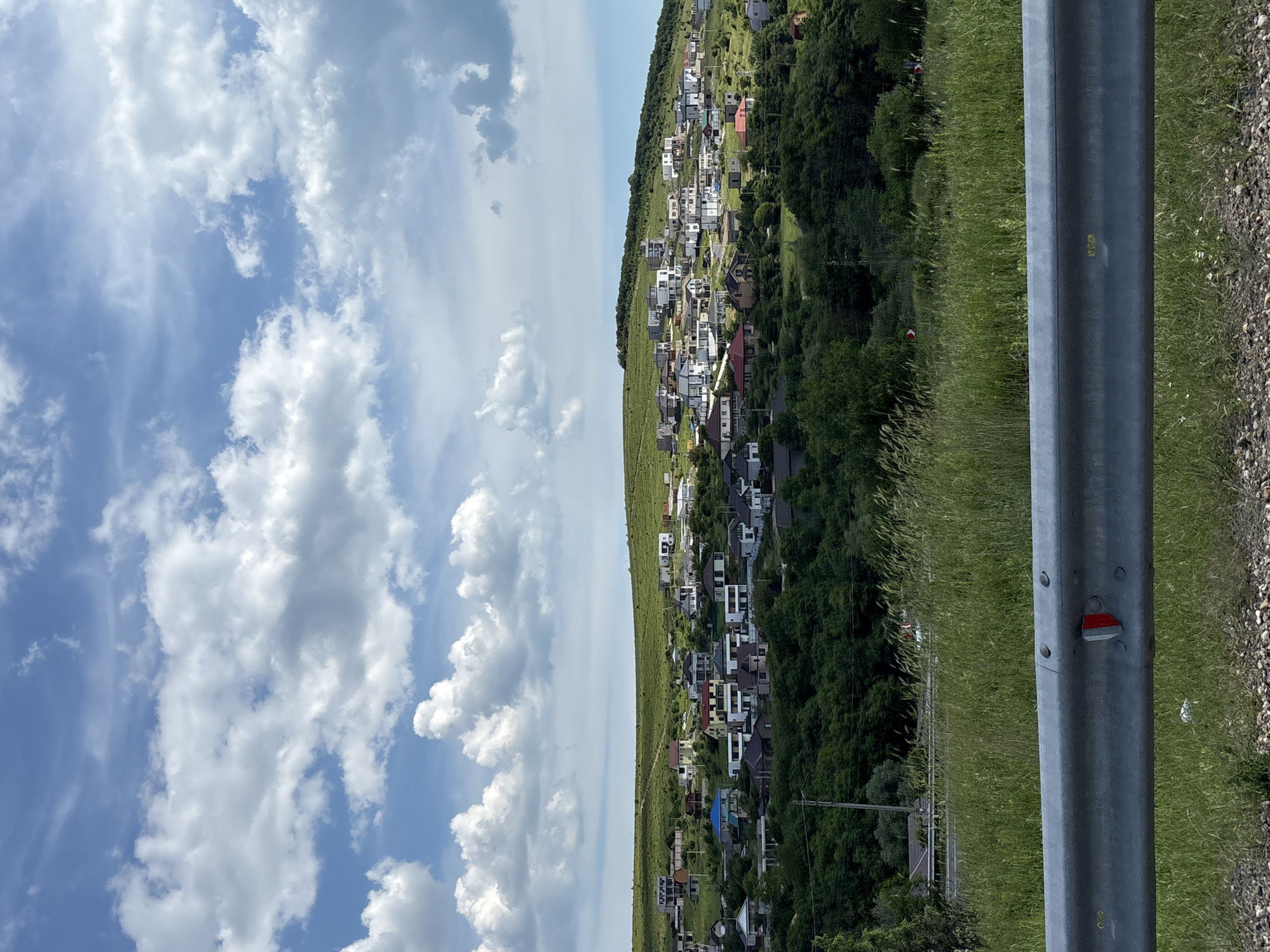 This image depicts a residential area situated on verdant, rolling hills in Varvarovka, Russia, under a partially cloudy blue sky during daylight hours. In the immediate foreground, a silver metal guardrail extends horizontally across the bottom of the frame, featuring a red and white reflector mounted near its center. Beneath and behind the guardrail, coarse gravel and tall green grass are visible. A dense band of dark green trees occupies the lower midground, separating the foreground from the cluster of houses. Numerous single and two-story houses are visible across the hillsides, showcasing varied architectural styles, some with white facades and modern designs, others with traditional colored roofs (red, blue, brown). The houses are interspersed with patches of green grass and foliage. The hills gently slope upwards, culminating in a relatively flat, green horizon line under a sky dominated by scattered, puffy white cumulus clouds with some lighter cirrus streaks. No individuals, specific actions, or interactions are discernible within the frame.