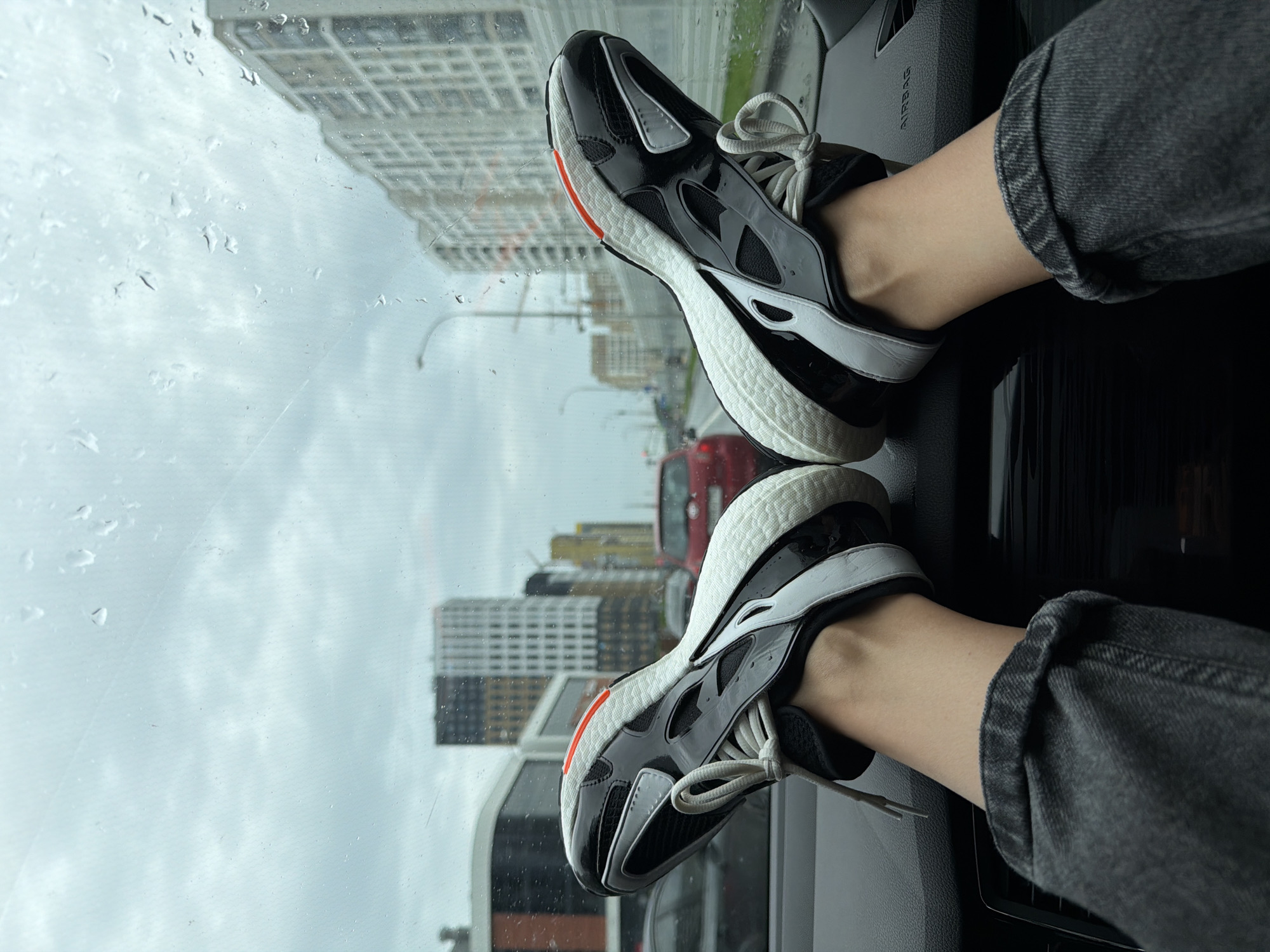 The image is an over-the-shoulder shot of a person's feet, clad in black and white athletic shoes with orange accents, resting on the dashboard of a car. The viewpoint is from inside the car, looking out through the windshield, which is speckled with raindrops. Outside, a city street is visible, with buildings, cars, and a lamp post under a cloudy, grey sky. The buildings are modern and vary in height and architectural style, with some featuring dark and light facades. A red car is in the middle ground, and other vehicles are further down the road. The dashboard is dark and textured, with a visible "AIRBAG" warning. The person is wearing dark, distressed denim jeans. The overall scene suggests a person is either driving or a passenger in a car, pausing on a rainy day in an urban environment.