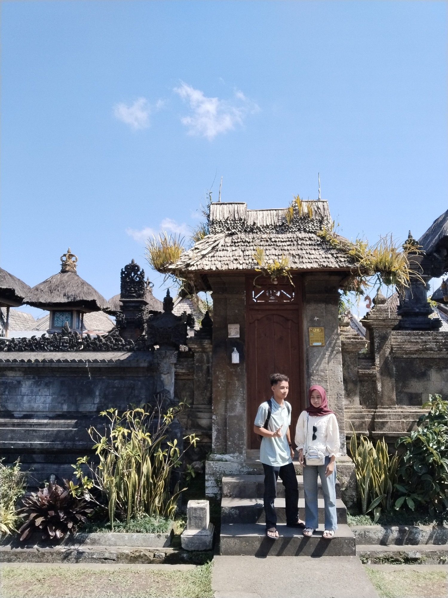 Two young people, a male and a female, stand on a stone staircase in front of a traditional Balinese temple entrance. The male, on the left, wears a light blue collared shirt, black pants, and sandals, with a backpack slung over his left shoulder. He is giving a peace sign with his right hand. The female, on the right, wears a white long-sleeved blouse, grey pants, and sandals, with a red hijab covering her hair. She has a small white crossbody bag.
The temple entrance features a weathered wooden door with intricate carvings and a tiered, thatched roof adorned with dry vegetation. The stone walls of the temple are dark and textured, decorated with ornamental carvings and statues. The surrounding environment includes lush green plants and trees, suggesting a tropical or subtropical climate. The sky is a clear, bright blue with a few scattered white clouds. The scene appears to be the entrance to a temple in Bangli, Indonesia, likely a place of cultural and religious significance.