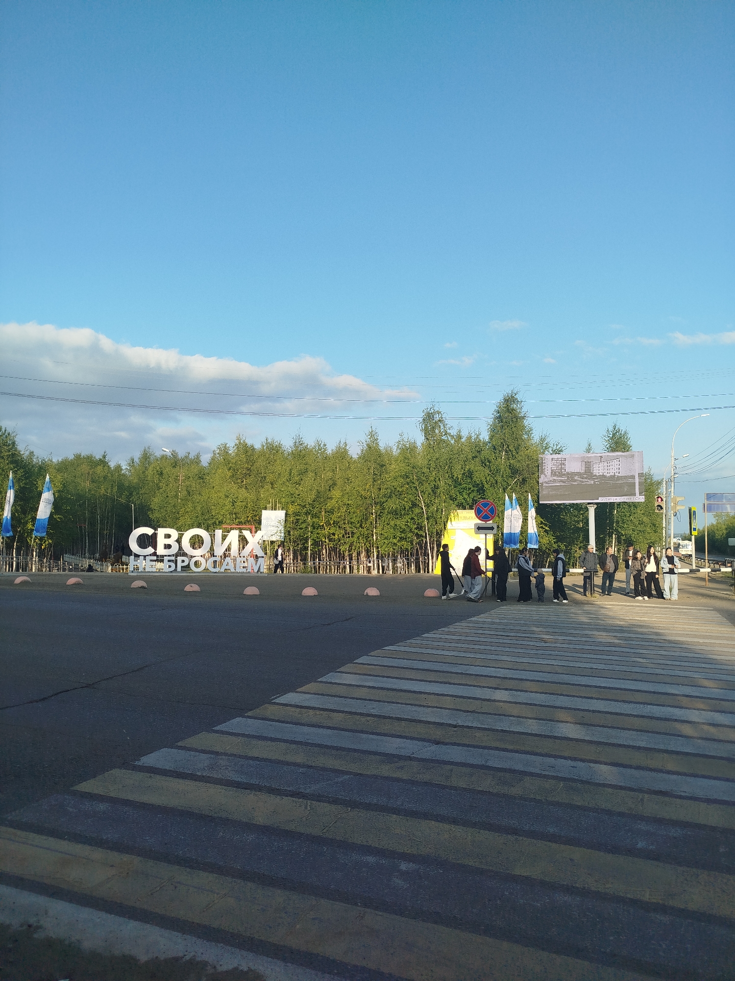 In the city of Mirny, Russia, a group of people are gathered at a pedestrian crossing. The scene is set against a backdrop of a lush green forest and a bright blue sky with scattered clouds. In the foreground, a large, white sculpture spelling out "СВОИХ НЕ БРОСАЕМ" (We do not abandon our own) is prominently displayed. Several flags, two of which are blue and white, are visible near the sculpture. A billboard featuring an architectural drawing is positioned behind the group of people. The pedestrian crossing itself is clearly marked with white and yellow stripes on the asphalt. Some individuals are standing, while others appear to be walking. The overall atmosphere suggests a public gathering or event taking place outdoors.