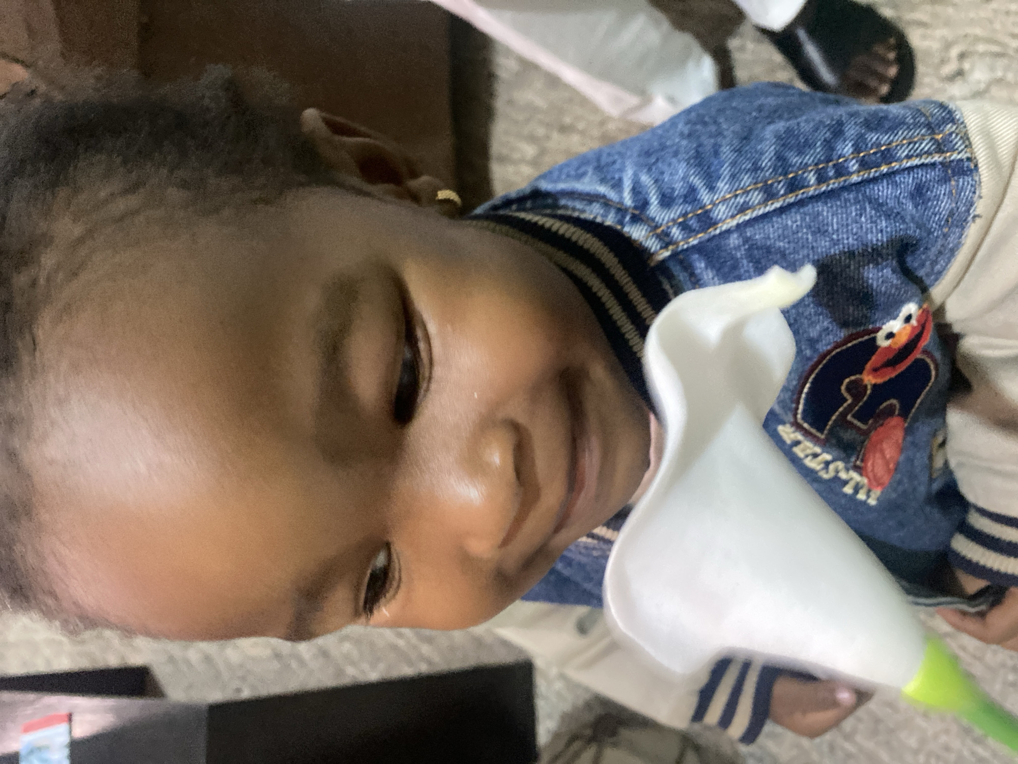 A young child with brown skin and dark hair holds a white calla lily flower. The child is wearing a denim jacket with a patch featuring a character, possibly Elmo from Sesame Street. The background is blurred, but there are glimpses of other people and flooring. The location in Bar Jirgi Summa, Nigeria is not evident from the image.
