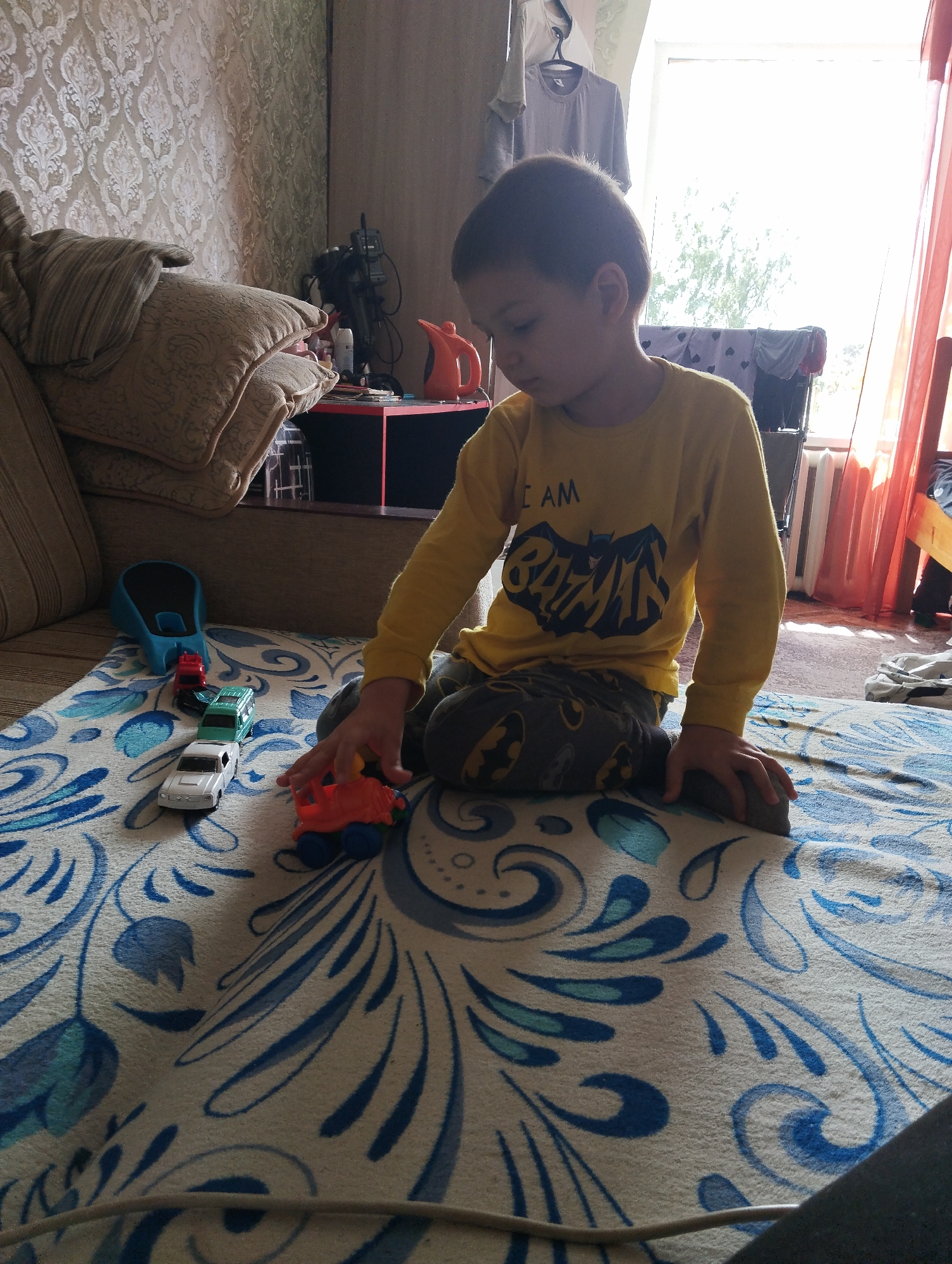A young boy wearing a yellow long-sleeved shirt with a Batman logo and dark pants with a Batman print is playing with toy cars on a patterned blanket. The blanket is white with blue and green floral designs. In front of the boy are three toy cars: a white car, a green car, and a red car, lined up in a row. The boy's right hand is reaching towards an orange and blue toy. To the left of the toy cars is a blue and black object. The scene appears to be indoors, possibly in a living room, with a couch visible in the background on the left. A window with light coming through is visible on the right, with curtains. The overall environment suggests a domestic setting. The location within the city of Kingisepp, Russia is not discernible from the visual information provided.