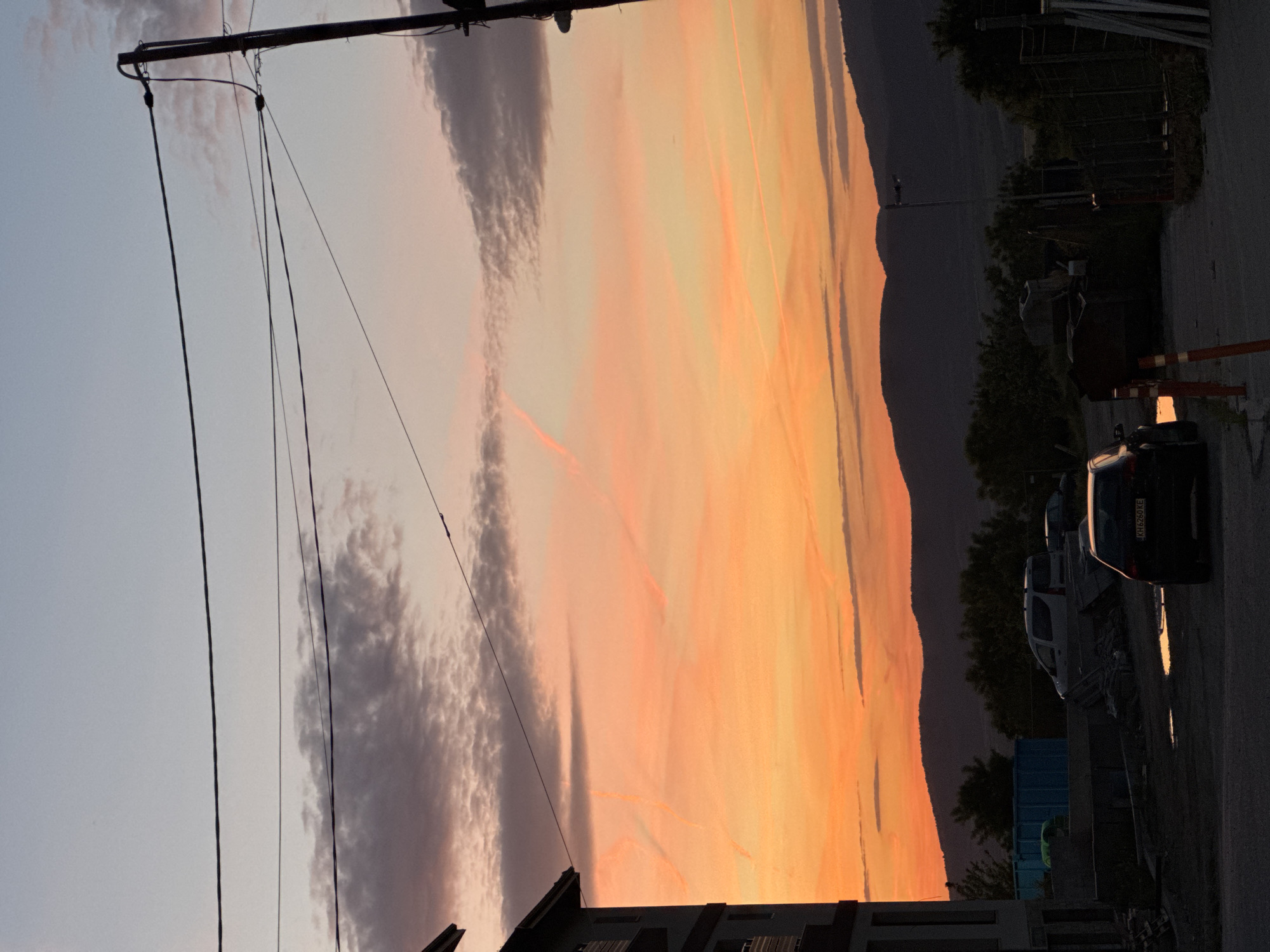 The image depicts a sunset in Sapareva Banya, Bulgaria. The sky is filled with orange and pink hues, contrasted by dark clouds. In the foreground, utility poles and wires are visible. The silhouette of a mountain range is present in the background. Parked vehicles and some buildings are visible in the lower portion of the frame.