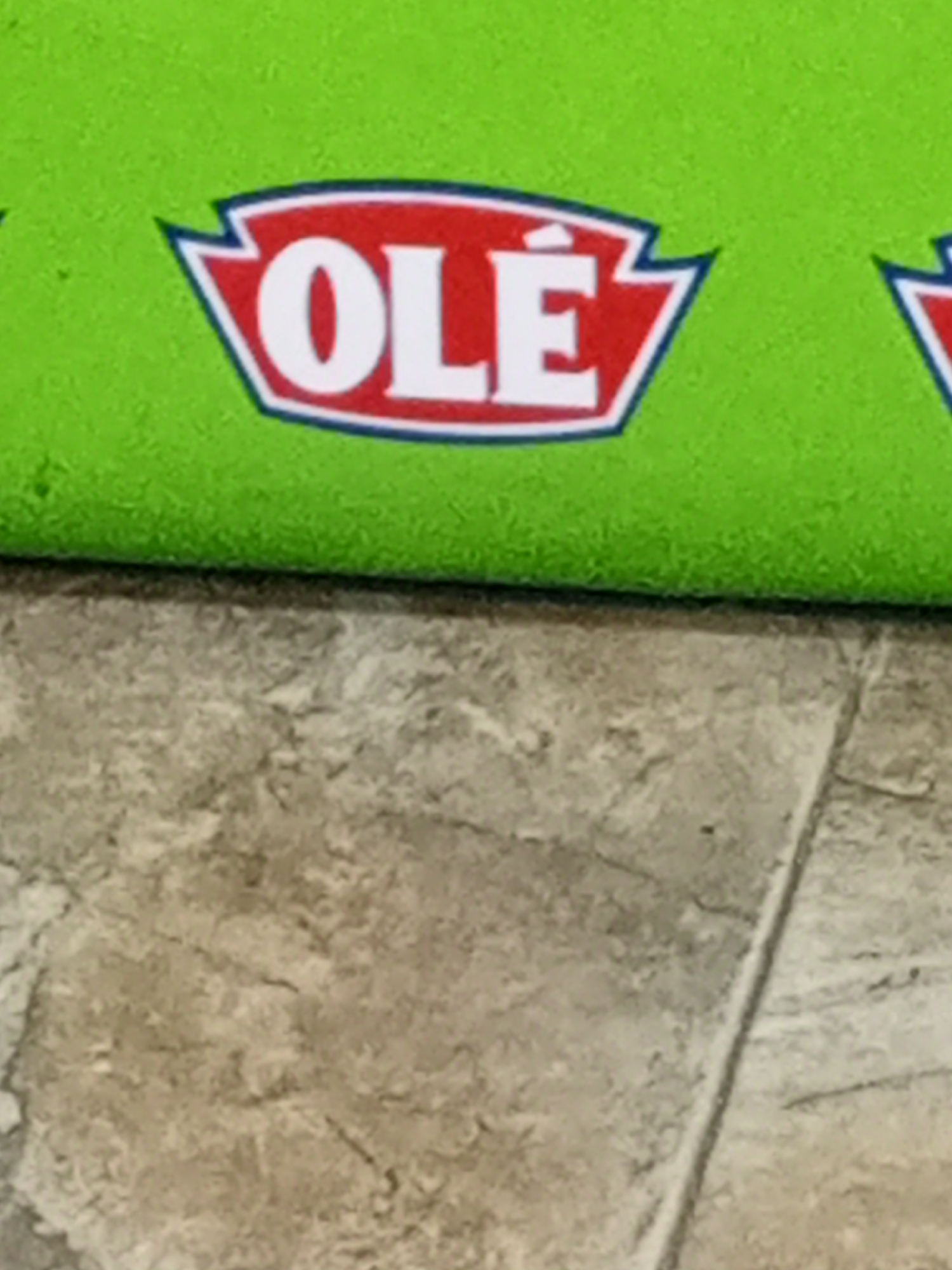 The image shows a green surface with the "OLÉ" logo, which is white text with a red outline in a pointed shape. The logo is partially visible, suggesting a larger product or packaging. The surface beneath the green object is a textured, light brown material, possibly tile or stone flooring. The scene provides no clues about a specific location in Juan Griego, Venezuela.