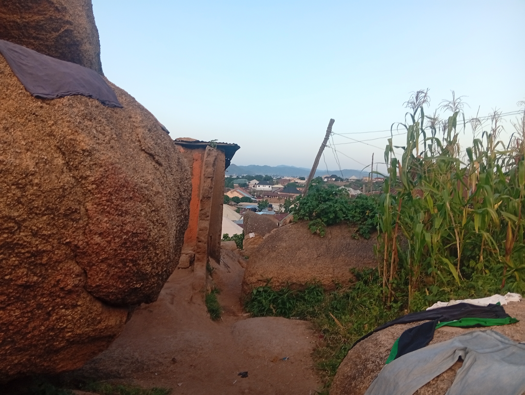 This image depicts an outdoor scene in Kabong, Nigeria, at what appears to be dusk or early morning given the soft lighting and clear sky. The foreground is dominated by large, weathered rocks, suggesting a natural or semi-natural landscape within or adjacent to the urban environment. On the largest rock in the left foreground, a dark blue piece of fabric is draped. A narrow, sandy path winds between these rocks, leading towards a more settled area in the midground.
The midground reveals a collection of buildings, characterized by corrugated metal roofs and what seem to be concrete or mud brick walls, indicative of typical residential structures in the region. A few trees and patches of green vegetation are interspersed among the buildings. To the right of the path, a row of tall corn stalks stands, with their feathery tassels reaching upwards. A wooden post with wires attached is visible among the corn, suggesting the presence of electricity or communication lines.
In the background, a hazy skyline of more buildings extends towards distant, low hills, faintly visible under the light sky. The overall impression is of a community nestled amongst a rocky terrain, with evidence of daily life, such as the drying fabric and agricultural elements like the corn. The environment appears somewhat arid, with dusty paths and sparse vegetation in some areas, contrasted by the lushness of the cornfield.