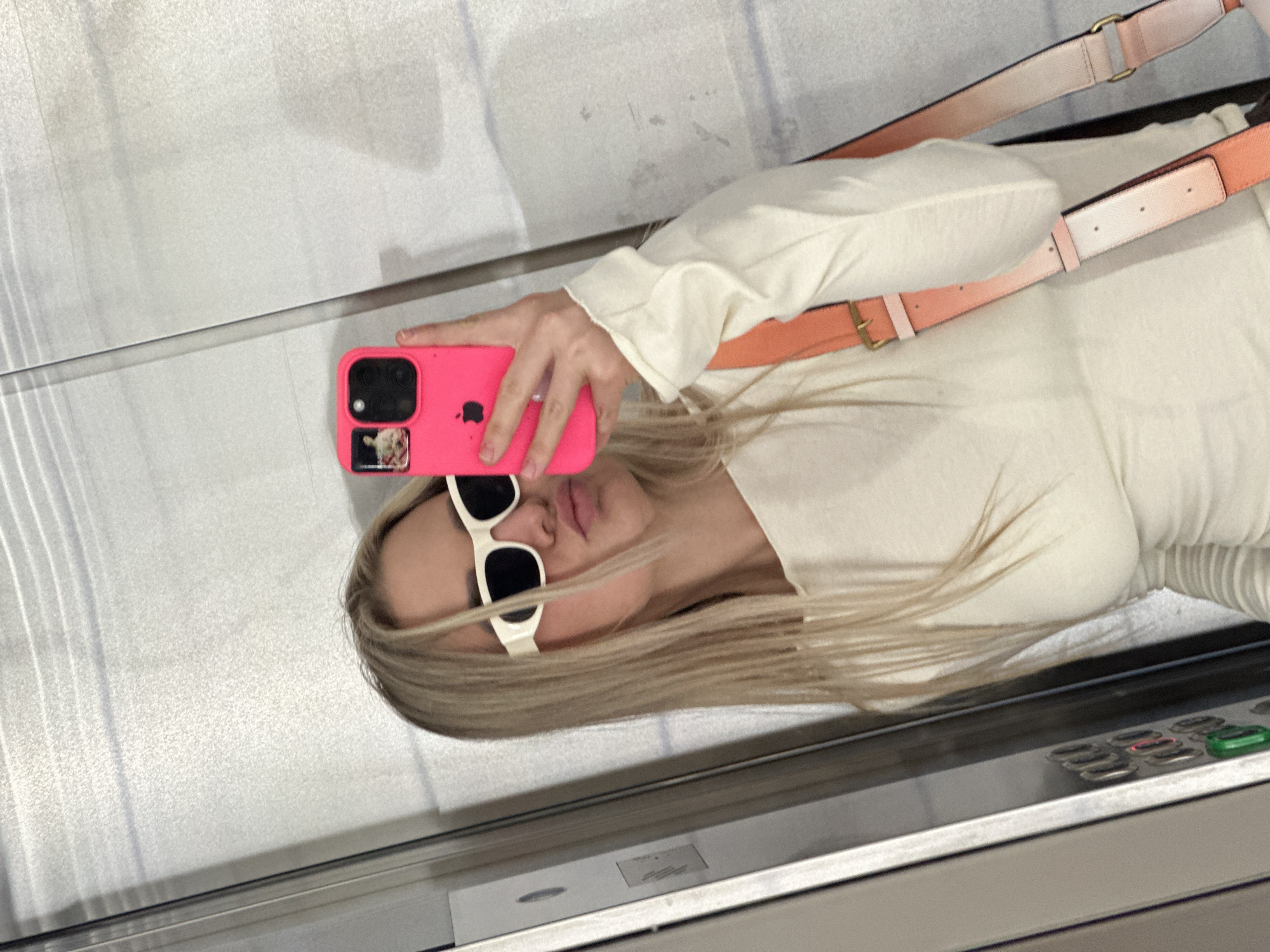 A woman with blonde hair is taking a selfie in an elevator. She is wearing white sunglasses and a light-colored long-sleeved shirt. The phone in her hand has a bright pink case. A purse strap is visible over her shoulder. The interior of the elevator is metallic. The image may be taken in Budva, Montenegro.