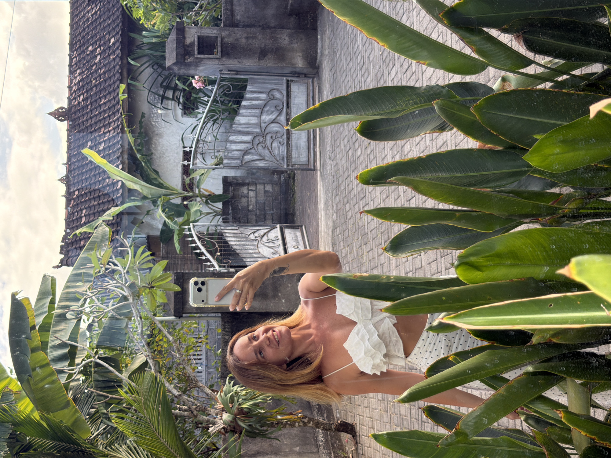 A woman with shoulder-length blonde hair takes a selfie with her smartphone in the city of Kuta Selatan, Indonesia. She is wearing a white ruffled crop top and white polka-dot pants. The woman is smiling and looking at her phone as she holds it up. In the background, a house with a tile roof and a decorative metal gate are visible. Lush green foliage, including large leaves, surrounds the woman. The ground is paved with cobblestones. The scene appears to be a residential area.