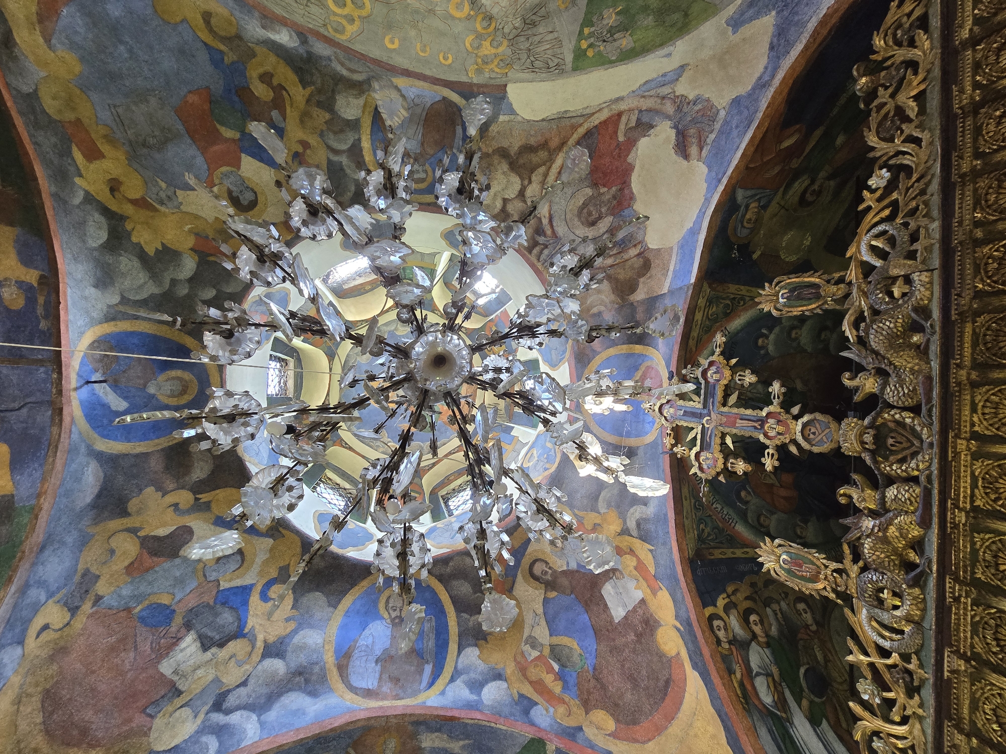 This image captures the interior of a church in Teteven, Bulgaria, presenting an upward view of a richly decorated domed ceiling and the upper portion of an ornate iconostasis.
A large, multi-tiered chandelier, composed of numerous clear crystalline or glass decorative elements and a metallic frame, hangs centrally from the apex of the domed ceiling. Light reflects off its facets, indicating ambient illumination.
The vaulted ceiling is extensively covered with frescoes depicting Christian iconography. These murals feature a palette of blues, reds, gold, browns, and muted greens. Circular medallions embedded within the design contain busts or full-figure representations of religious figures, including individuals holding books, consistent with evangelists or saints. Surrounding these medallions are stylized clouds, decorative scrolling patterns in gold, and depictions of angels. The artistic style is characteristic of Orthodox iconography. Portions of the murals show signs of age and wear, including some discoloration and minor flaking.
At the bottom of the frame, the upper section of an elaborate, carved and gilded wooden iconostasis is visible. This structure features intricate carvings, including serpentine or dragon-like figures flanking a central element, interspersed with floral and foliate motifs. Mounted atop the iconostasis is a large, ornate crucifix depicting Jesus Christ on the cross, flanked by smaller icons or decorative elements. Additional framed icons are positioned on the iconostasis, one showing a standing saint. Behind the iconostasis, further murals are discernible on the wall or a lower section of the ceiling, portraying groups of figures, likely saints or congregants.