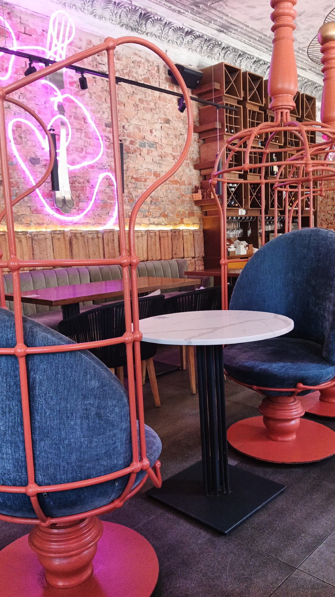 The image depicts the interior of a cafe or restaurant in Kryvyi Rih, Ukraine. The seating area features unique chairs with red metal cage-like frames enclosing dark blue upholstered seats. These chairs are arranged around small, round tables with white marble-like tops and black metal bases. In the background, a brick wall is visible, adorned with a vibrant pink neon light installation that appears to be abstract or stylized text. Further back, there are more seating areas with upholstered benches and dark chairs, as well as wooden tables. A wall-mounted wine rack filled with bottles is also visible in the background. The overall ambiance suggests a modern and stylish establishment.