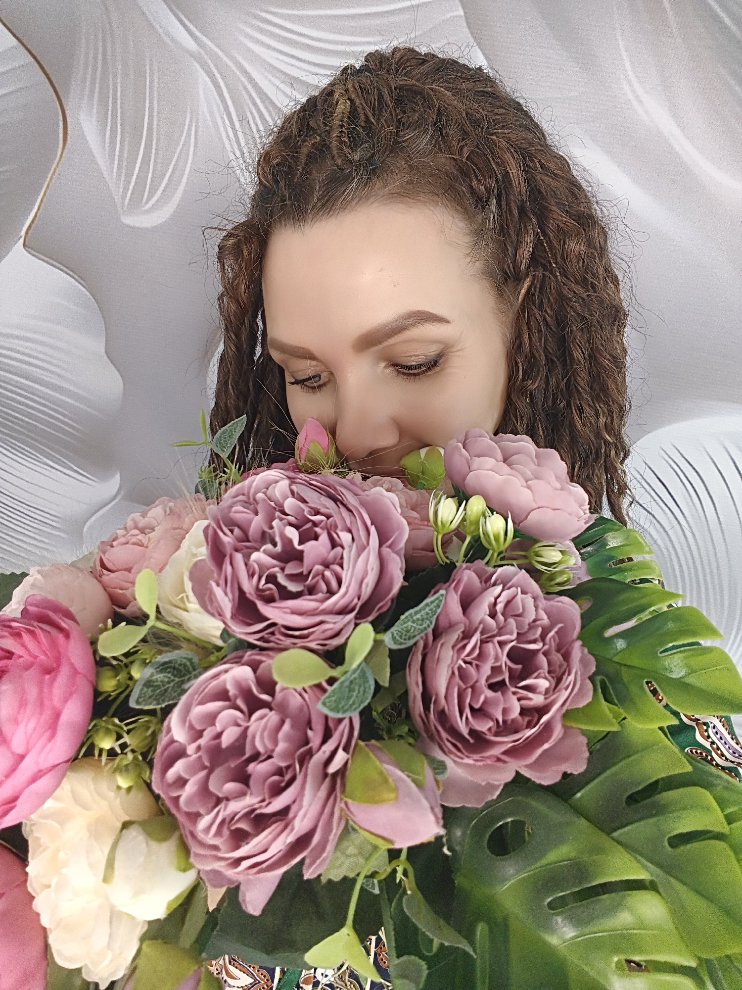 A woman with curly brown hair, styled with some strands braided near the forehead, is seen in a close-up shot smelling a bouquet of flowers. The flowers are a mix of pink, off-white, and mauve roses, along with some green buds and foliage, including large, slotted tropical leaves. The background is a white, textured surface with curved lines, possibly a decorative wall panel. The scene appears to be indoors. The city and specific location are unknown.