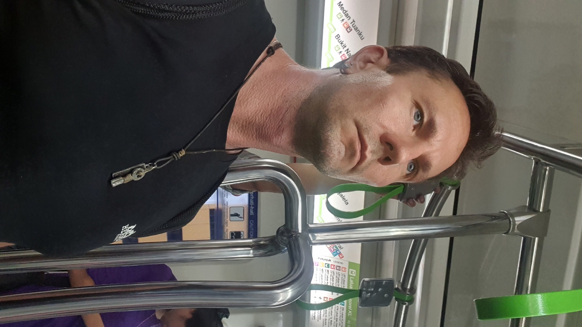 A middle-aged Caucasian man with short brown hair and blue eyes is the primary subject of this medium close-up, taken from a low angle. He is wearing a black t-shirt and a silver earring in his left ear. A dark cord necklace with a silver key pendant is visible around his neck. The man appears to be inside a public transportation vehicle, likely a train, in Kuala Lumpur, Malaysia. Signs in Malay are visible in the background, including "Medan Tuanku" and "Bukit Nanas," which are stations on the Kelana Jaya LRT line. Green hand straps are hanging from the ceiling, and metal poles are present, typical of a train interior. Another person, partially obscured and wearing a purple shirt, is visible in the background on the right. The lighting suggests an indoor environment, possibly during the daytime.
