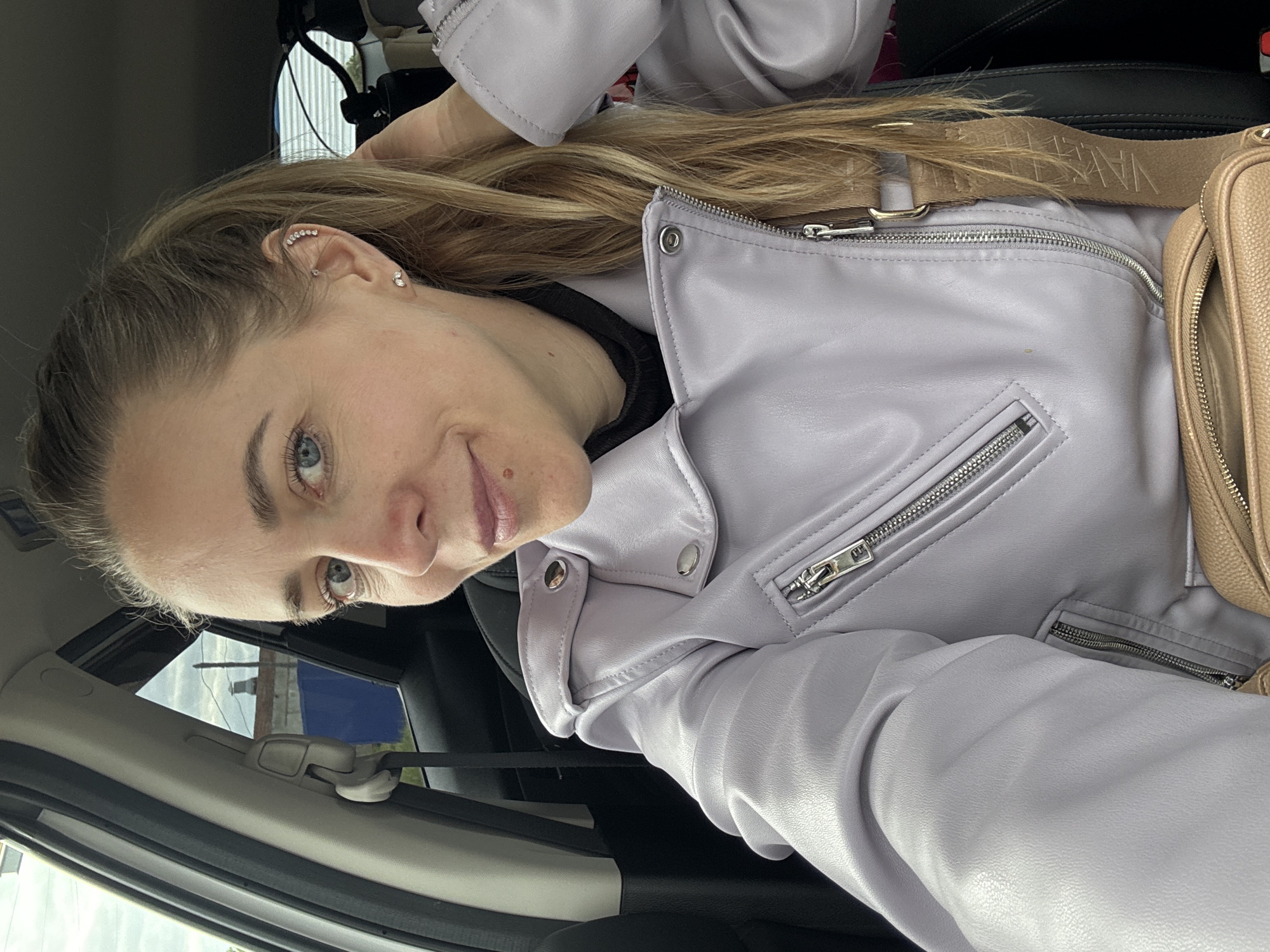 A woman with light skin and blue eyes is taking a selfie in what appears to be a car. She has blond hair pulled back, revealing ear piercings. She is wearing a light purple leather jacket. A tan purse with a strap is visible over her shoulder. The interior of the car includes a black seat and trim, and a beige headliner. The location is not clear, and thus not able to be confirmed as Chaikovsky, Russia.