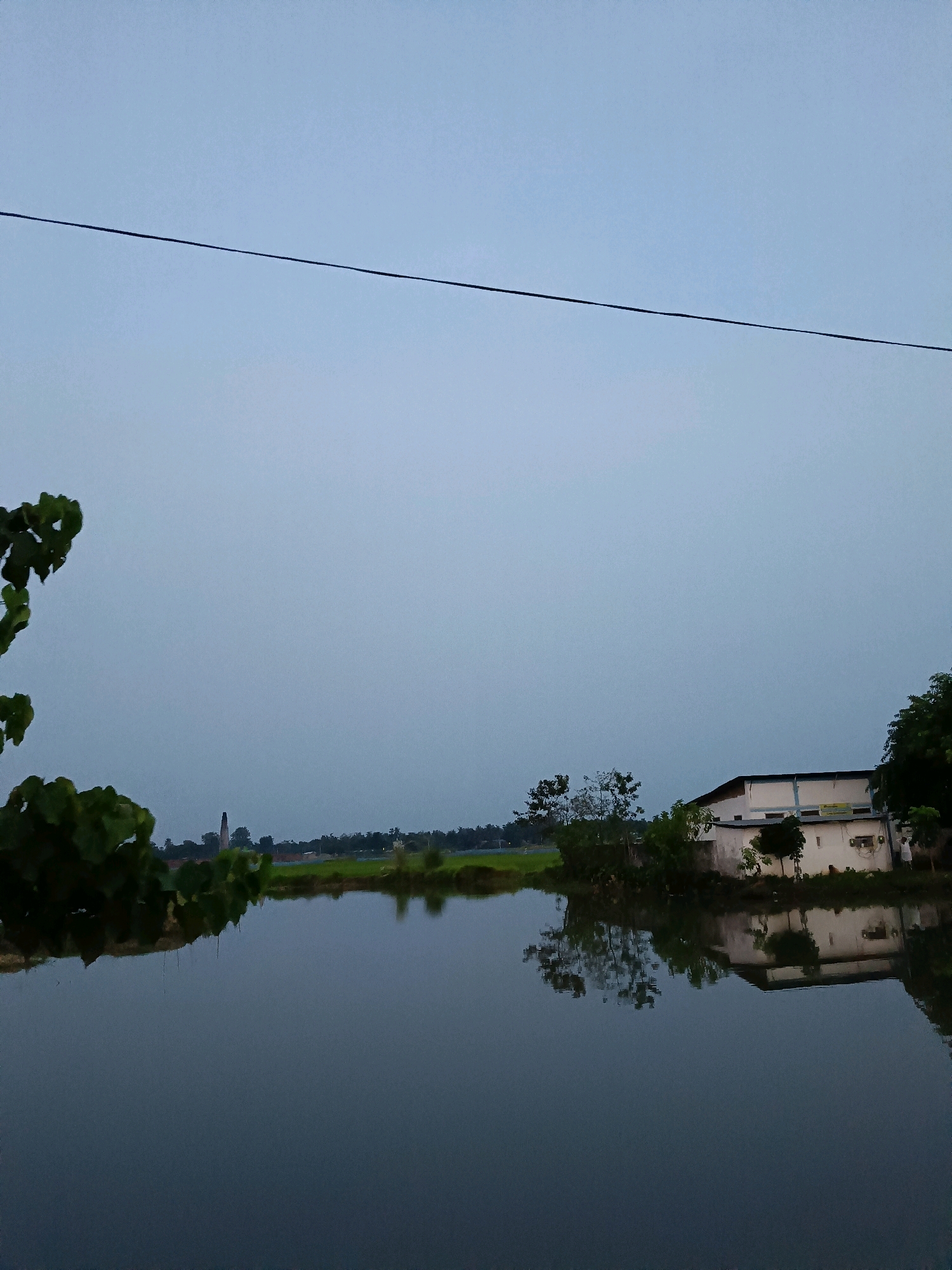 The image depicts a serene natural scene, possibly at dusk or dawn, featuring a calm body of water reflecting the sky and surrounding environment. In the foreground, a portion of lush green foliage is visible on the left, partially obscuring the view. The water stretches across the lower half of the image, smooth and dark, creating a mirror-like surface.
In the middle ground, a low-lying landscape is visible, with patches of greenery and a distant, tall, conical structure that resembles a brick kiln, a common sight in rural industrial areas. Further to the right, a modern building with a white facade and blue trim is situated, partially obscured by trees. This building appears to be a residential or commercial establishment. Trees with dense canopies are scattered throughout the landscape, adding to the natural ambiance. The reflection of the building and trees is clearly visible on the water's surface.
The sky above is a pale, overcast blue, suggesting subdued lighting conditions. A thin, dark electrical wire stretches horizontally across the upper portion of the frame, dividing the sky. There are no visible people in the image, and no discernible actions or interactions are taking place. The overall atmosphere is one of tranquility and stillness.
Based on the characteristic architecture of the building, the presence of a brick kiln, and the surrounding agricultural landscape, this scene is likely located in or near the city of Barpeta, India, a region known for its rural character and traditional industries.
