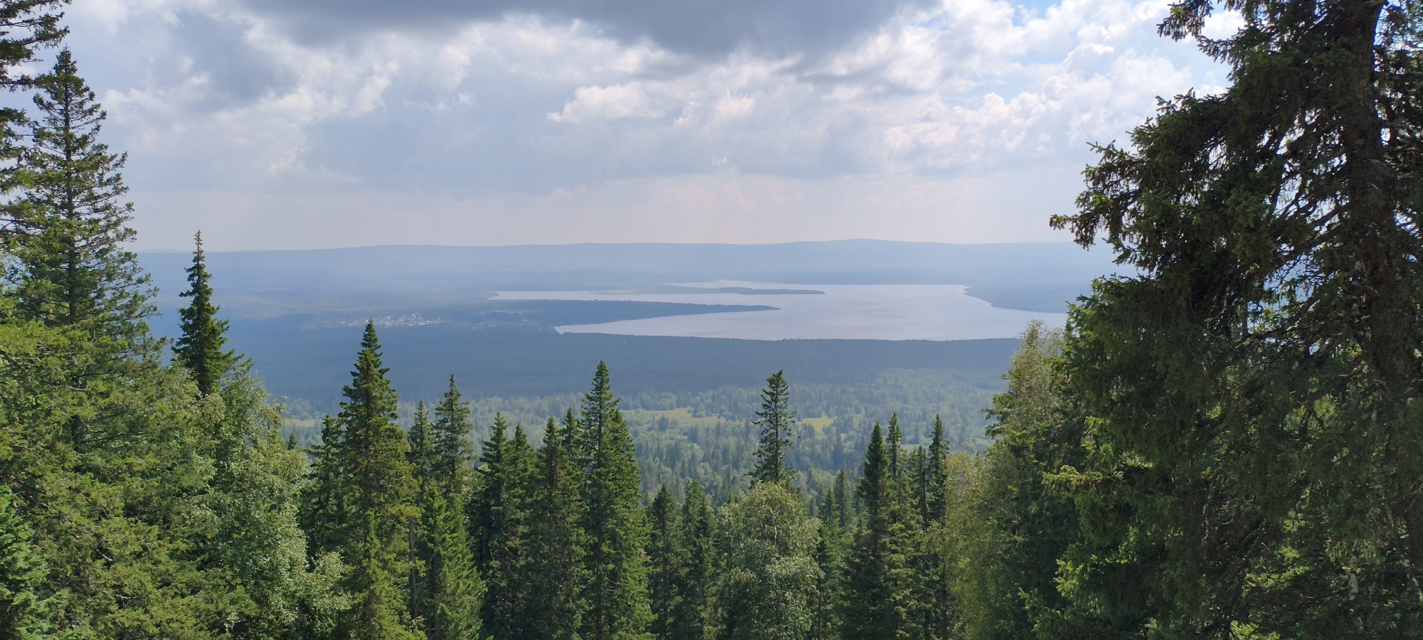 This is a panoramic, high-angle landscape photograph of a mountainous region, taken from a forested vantage point overlooking a large body of water and a vast expanse of forest. The foreground is dominated by the dense canopy of coniferous trees, primarily fir and pine, with a few deciduous trees interspersed. The trees are a deep green, with many reaching towards the sky.
In the midground, a wide, calm body of water, likely Lake Ziuratkul', stretches across the horizon. The lake's surface is a muted blue-grey, reflecting the overcast sky. Along the shoreline of the lake, a dark, dense forest can be seen, transitioning into lighter green patches of vegetation further inland. A small cluster of buildings, indicative of a settlement, is visible on the far left edge of the lake.
The background consists of a hazy, flat horizon line, with the distant mountains appearing as soft, blue-grey silhouettes against the sky. The sky itself is partly cloudy, with large, dramatic grey clouds interspersed with patches of bright white. The lighting suggests a bright, possibly late afternoon, but diffused by the cloud cover.
The location is identified as Ziuratkul', Russia, likely a scenic overlook or a high point within the national park, offering a commanding view of the lake and surrounding wilderness. There are no people or animals visible in the image, and no discernible actions or interactions are taking place. The scene is static and depicts a natural, undeveloped landscape.