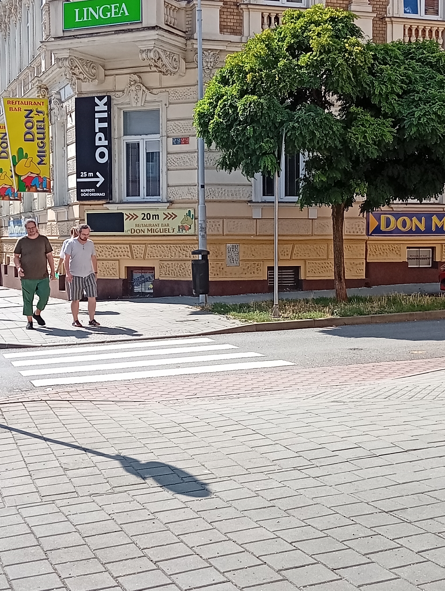 The image depicts a street scene in Brno, Czechia, focusing on a building with multiple signs and a crosswalk. On the facade of the building, a green sign reads "LINGEA". To the left, a yellow banner displays "RESTAURANT BAR DON MIGUEL" with cartoon depictions. A black vertical sign with white text says "OPTIK" and points to the right with "25 m NAPROTI OČNÍ ORDINACI". Below this, another yellow sign reads "20 m RESTAURANT BAR DON MIGUEL'S". Two men are walking across a crosswalk in the foreground. The left man wears a dark t-shirt and green pants, while the right man wears a light t-shirt and striped shorts. A tree with lush green foliage is situated to the right of the building. The street is paved with interlocking bricks, and the crosswalk is clearly marked with white stripes. The overall environment appears to be a commercial area within a city.