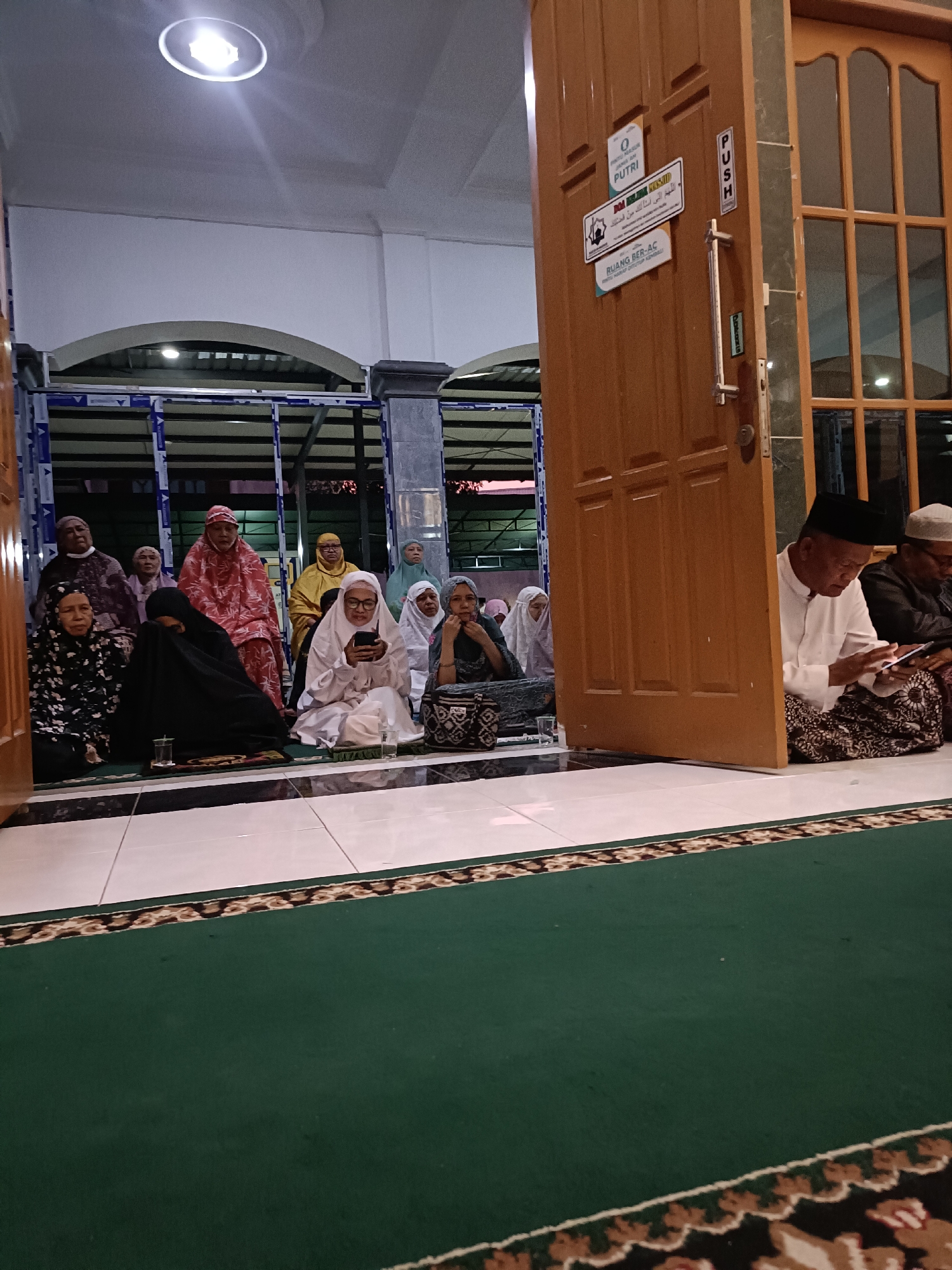 The image shows a group of people, predominantly women, sitting on a green carpet inside what appears to be a mosque or prayer hall in the city of Kediri, Indonesia. The lighting suggests it is evening or nighttime.
In the foreground, partially obscured by a wooden door, an elderly man wearing a white shirt and a peci (a traditional cap) is looking at a mobile phone. To his right, another man, also wearing a peci and traditional attire, is visible.
The main group of people consists of at least ten individuals, all appearing to be adults, seated on the floor. Most of them are women, dressed in various modest Islamic attire, including hijabs and long garments. Some are looking forward, while others are engaged in their own activities, such as one woman holding a mobile phone and another with a book. Two glasses of water are visible on the floor.
The environment includes a tiled floor, a green carpet with a decorative border, and a large wooden door with glass panels. The door has several signs attached to it, one of which reads "PUSTRI" (likely indicating "Putri" which means "daughter" or "female" in Indonesian, suggesting a women's section or facility) and another with Arabic script. A "PUSH" sign is also visible on the door. In the background, architectural elements of the building, including arches and a ceiling with a bright light fixture, are discernible. The overall impression is a serene gathering for prayer or religious activity.