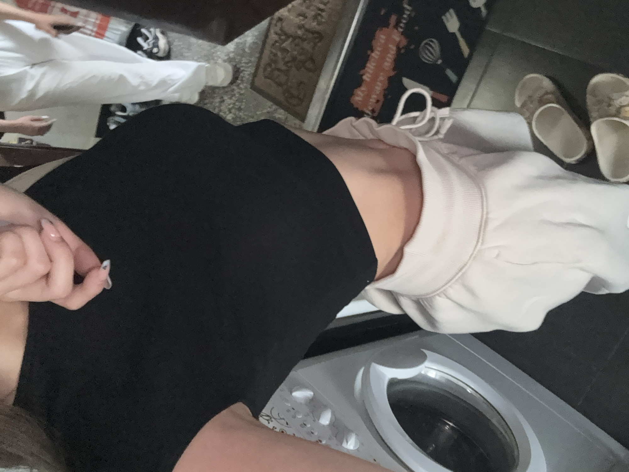 A person is standing in front of a washing machine in what appears to be a home environment. The person is wearing a black crop top and light-colored sweatpants. They are holding up their hand, showing off their nails. A pair of sandals is visible on the floor. The background includes a doormat with text on it. There is no discernible indication that this scene is located in Sofia, Bulgaria.