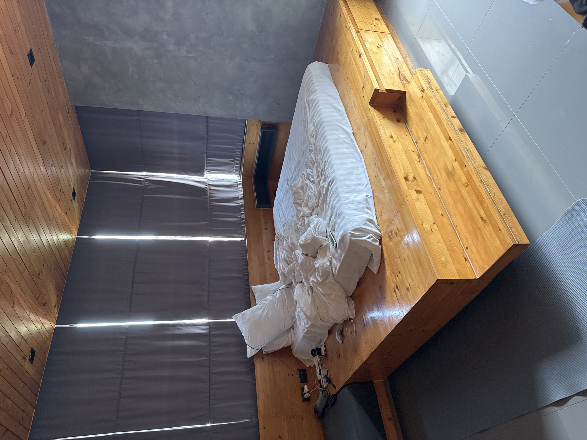 This image depicts a bedroom in Rawai, Thailand, with a large, low-profile bed made of light-colored wood. The bed is dressed in rumpled white bedding with faint vertical stripes, and two pillows are propped at the head. To the left of the bed, a dark gray upholstered platform serves as a seating area or bedside extension, with a green water bottle and various electronic devices, including chargers and a remote control, on the wooden surface. The headboard is also made of wood and integrates into the wall. A dark gray, textured wall is visible on the right side of the room. The ceiling is paneled with warm-toned wood, and a dark gray curtain covers the windows, allowing vertical shafts of light to enter. The floor is tiled in a light gray color. A dark gray yoga mat is placed on the floor in front of the wooden bed platform. The overall impression is of a modern, minimalist bedroom with natural wood accents.