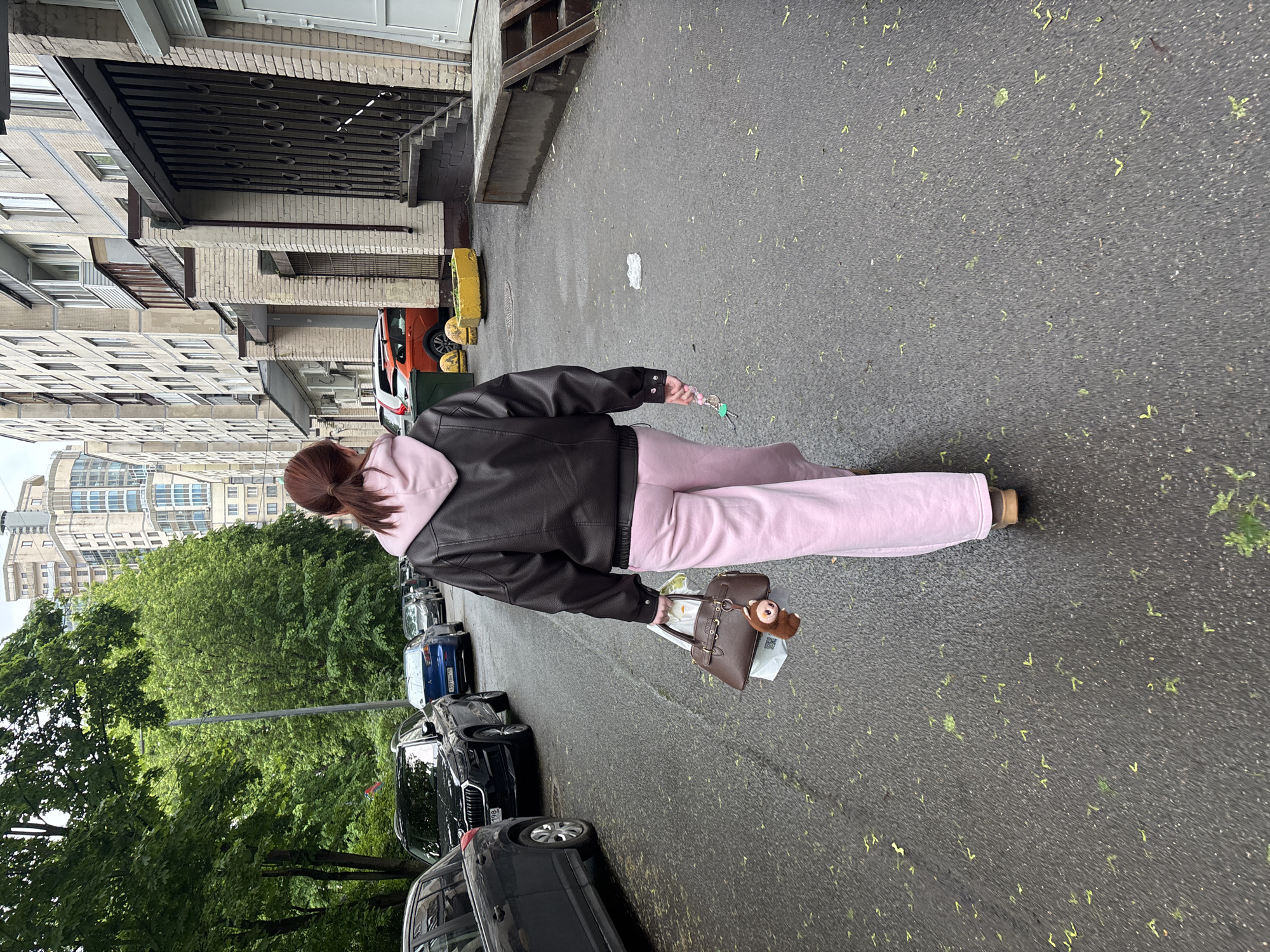 A woman walks on a wet asphalt street lined with parked cars and buildings. She is wearing a black jacket over a light pink hoodie, light pink pants, and has her reddish-brown hair pulled back in a ponytail. She carries a brown purse with a small monkey keychain attached. The street is marked with scattered green debris. Buildings of varying architectural styles are visible on either side of the street. One building features a decorative metal grate above the entrance. In the background, dense green trees are visible, obscuring parts of additional buildings. Based on architectural style and the license plates on the cars, the location is likely in Saint Petersburg, Russia.