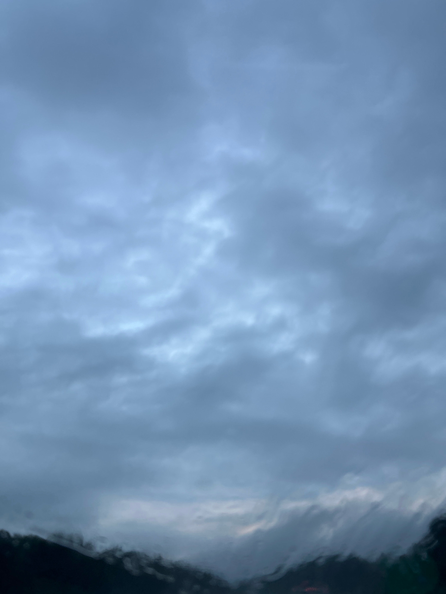 The image displays a low-angle view of a cloudy sky, predominantly gray and blue hues, with some areas appearing lighter, possibly due to breaks in the cloud cover. The bottom portion of the image is obscured by dark, blurry shapes that suggest a landscape or city skyline, rendered indistinct by what appears to be rain or moisture on a viewing surface, such as a car windshield. No people, specific objects, or discernible actions are visible. The overall impression is one of overcast weather, potentially during or after rainfall. The location is identified as Collier Township, United States of America.