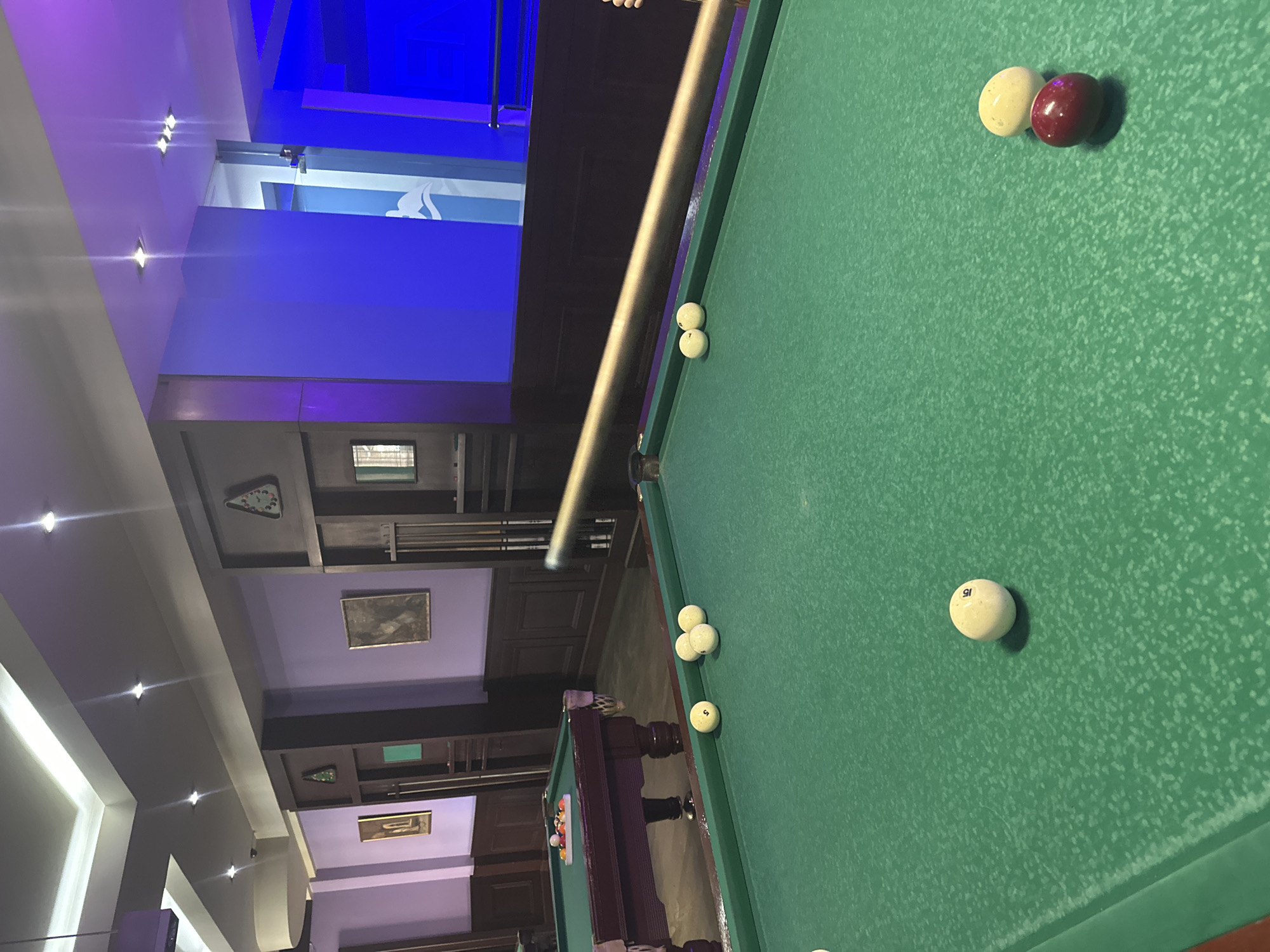 A billiard table is in a billiards room. The table is covered in green felt and has several billiard balls on it. One is white with the number 15 on it. Another is white. There is also a red billiard ball. A cue stick is positioned in front of the ball. The billiards room has purple walls, recessed lighting, and paintings. There are cue racks on the wall.