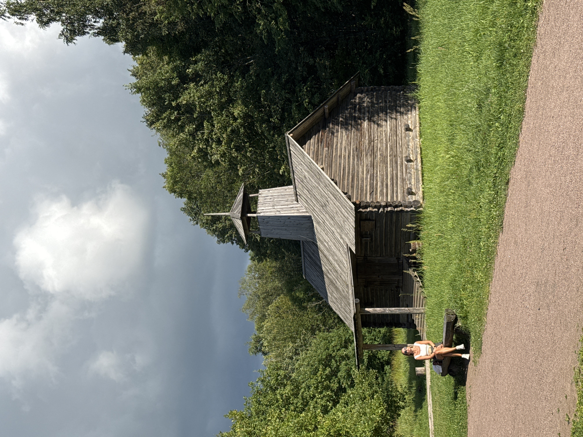 A woman is seated on a wooden bench in front of a rustic wooden building in an outdoor setting. The building appears to be historical, with a thatched roof and a small tower structure. The woman is wearing a white tank top and a skirt, with white sneakers. She has on sunglasses and is looking towards the right of the frame. She is positioned on the left side of the image. The building is in the center and right of the image. The ground in front of the building is covered in lush green grass, with a pathway made of gravel extending from the bottom left corner of the image towards the building. Trees and foliage form a dense backdrop behind the building. The sky above is partly cloudy with dramatic, dark clouds on the left side and brighter clouds on the right. The overall scene suggests a park or historical park setting in Veliky Novgorod, Russia, possibly depicting a traditional structure.